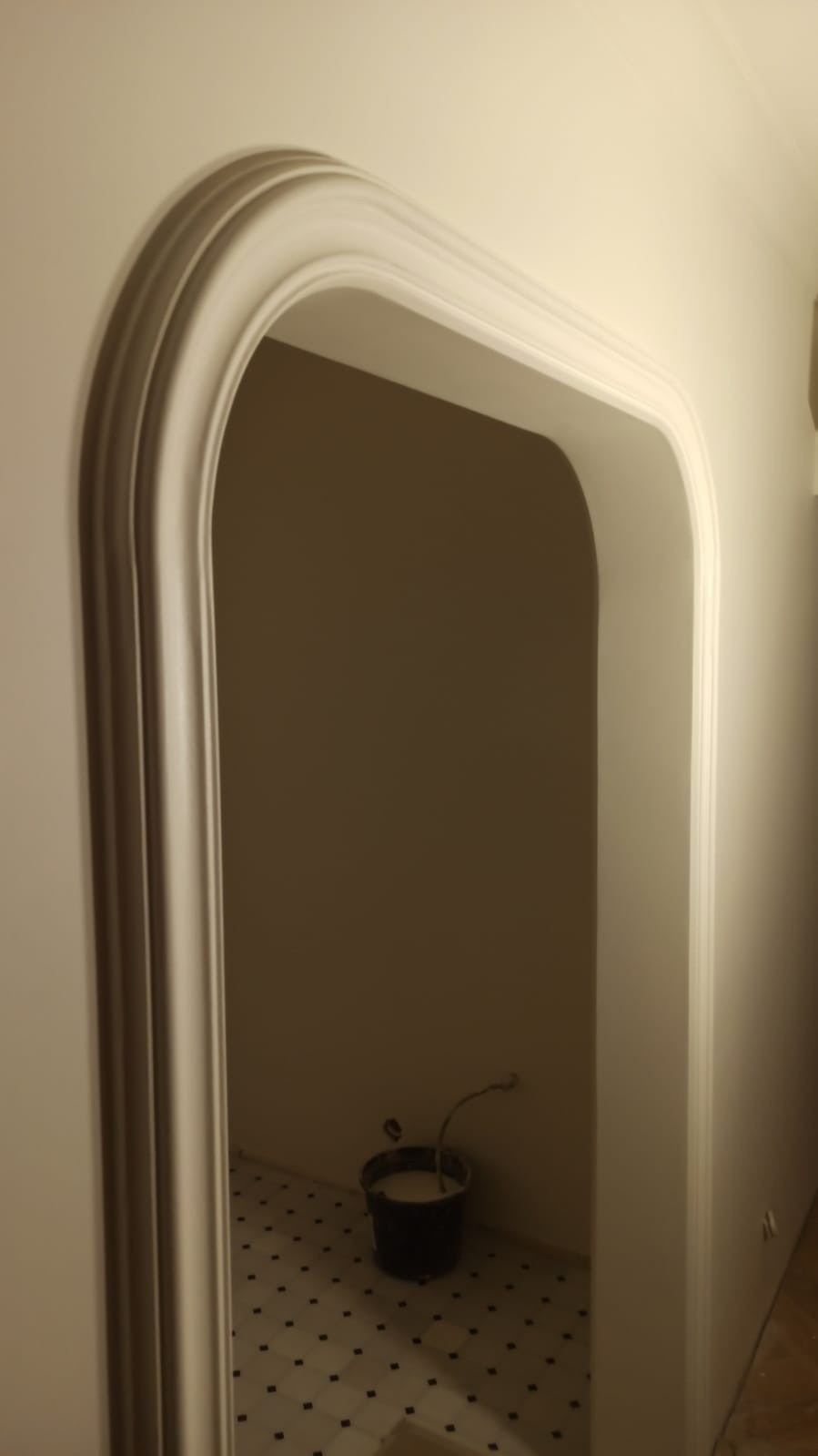 Biały, zaokrąglony portal drzwiowy z dekoracyjnymi listwami, widok na wnętrze z wiadrem i wzorzystą podłogą. Świeżo pomalowane ściany w neutralnych odcieniach.