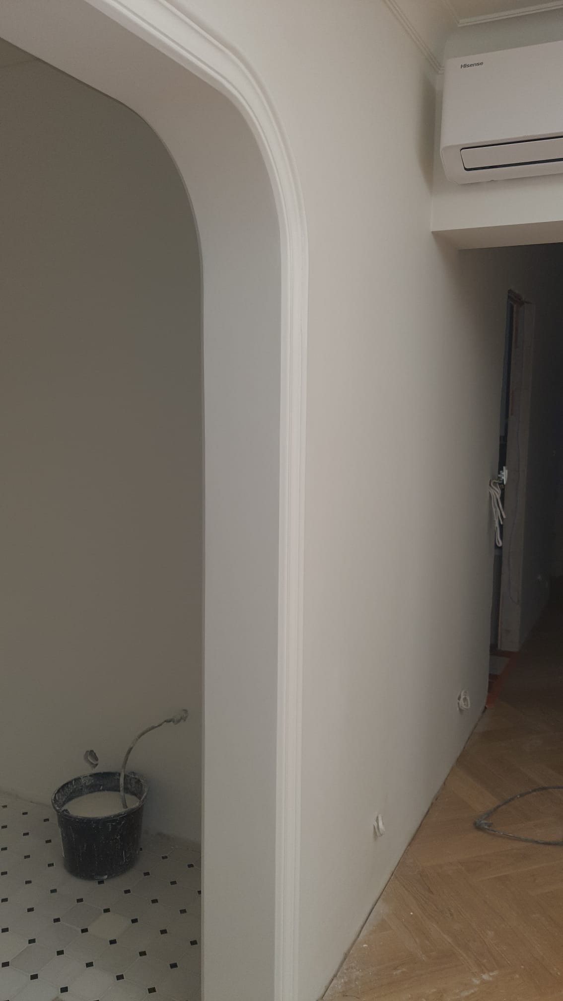 Białe, świeżo pomalowane wnętrze z łukowym przejściem i sztukaterią. Widoczny fragment podłogi w szachownicę i wiadro z farbą. Minimalistyczny design.