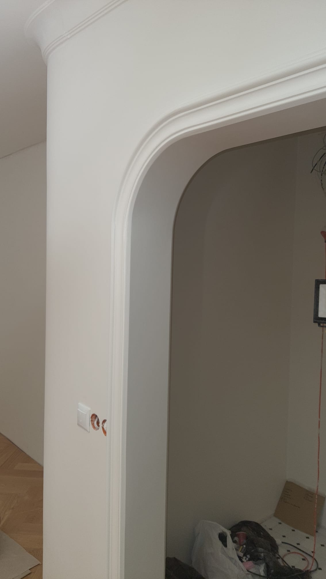 Białe, wykończone przejście łukowe z dekoracyjną sztukaterią i gładkimi ścianami. Widoczne gniazdka elektryczne bez osprzętu i fragment parkietu.
