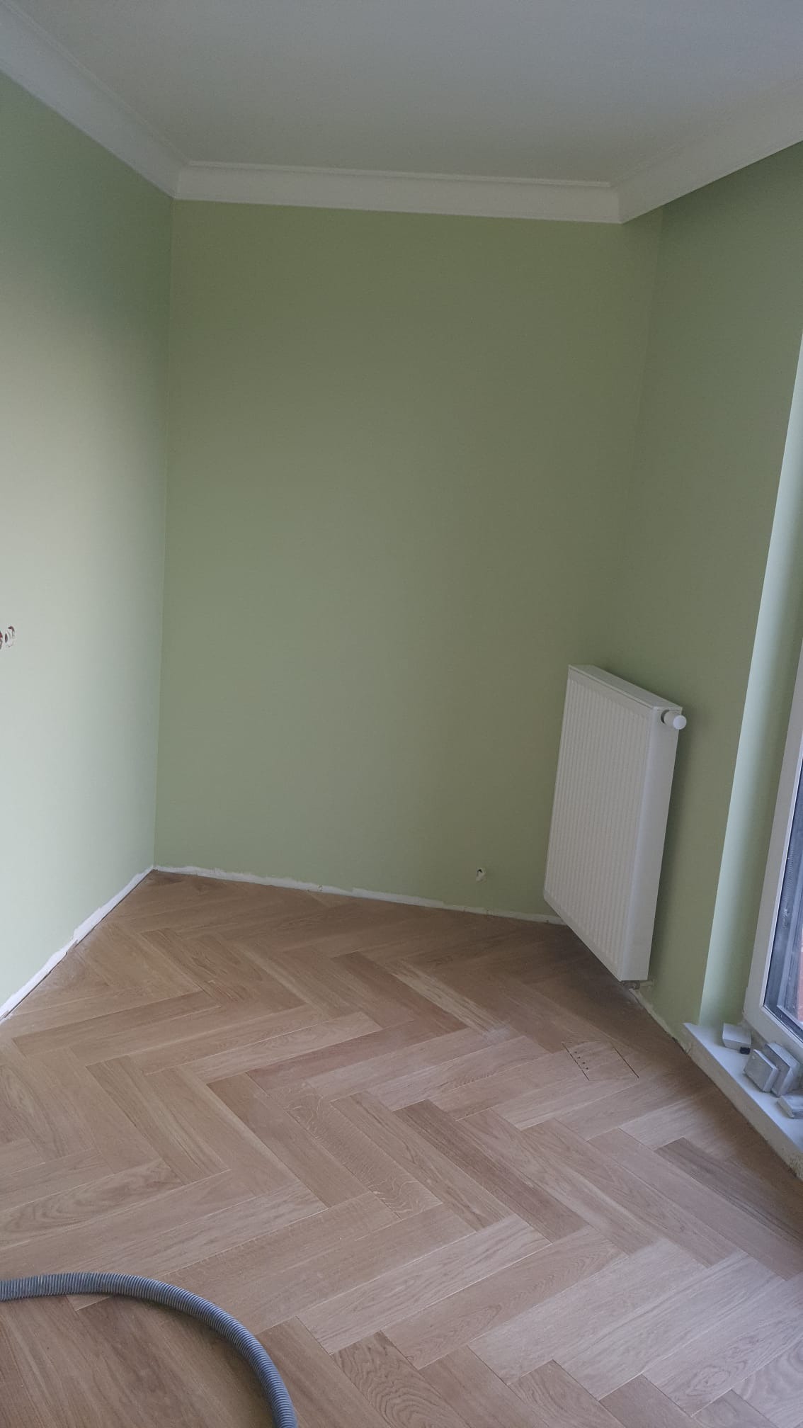 Pokój po remoncie: parkiet w jodełkę, świeżo pomalowane ściany na zielono, biały grzejnik i listwy przypodłogowe. Widoczny fragment węża od odkurzacza.