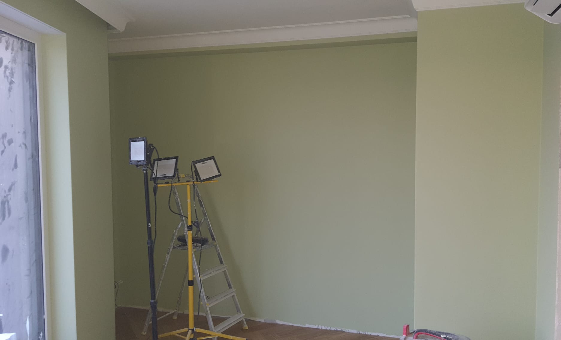 Pomalowane na jasnozielono ściany w pokoju, drabina malarska z reflektorami, widoczne listwy przypodłogowe i fragment okna. Świeżo po malowaniu.