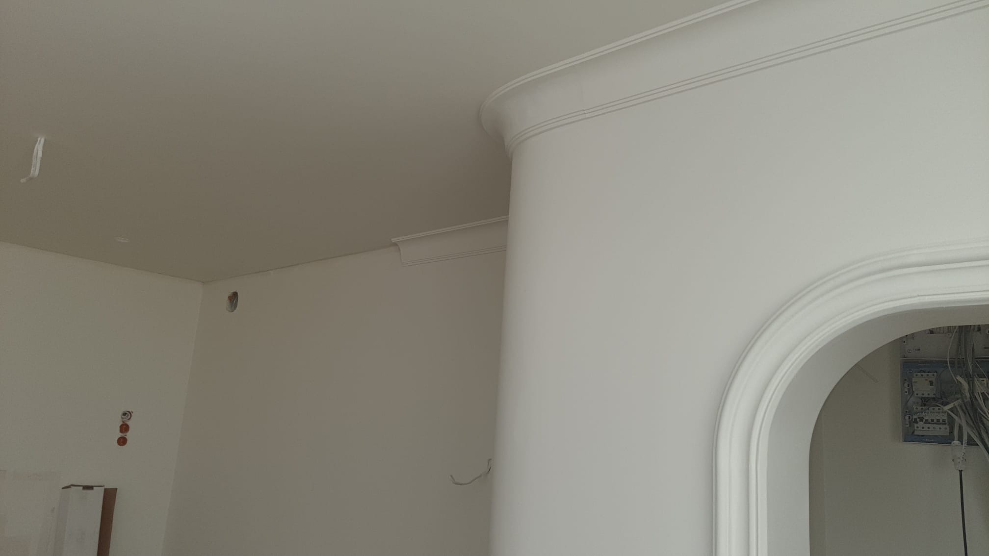 Białe ściany z dekoracyjnymi listwami przysufitowymi i łukowym przejściem. Widoczne punkty elektryczne i przewody. Minimalistyczne wykończenie wnętrza.