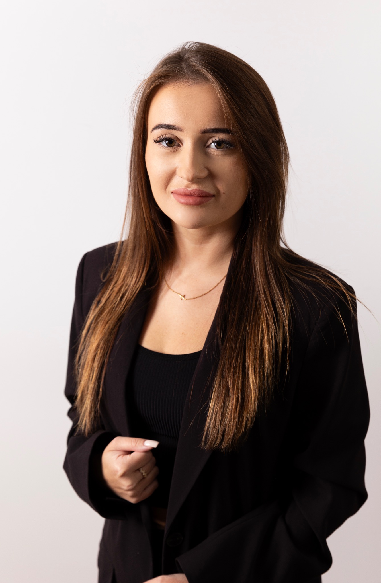 Portret kobiety w czarnej marynarce i bluzce, z delikatnym makijażem i długimi brązowymi włosami, na jasnym tle. Subtelna biżuteria podkreśla elegancję.