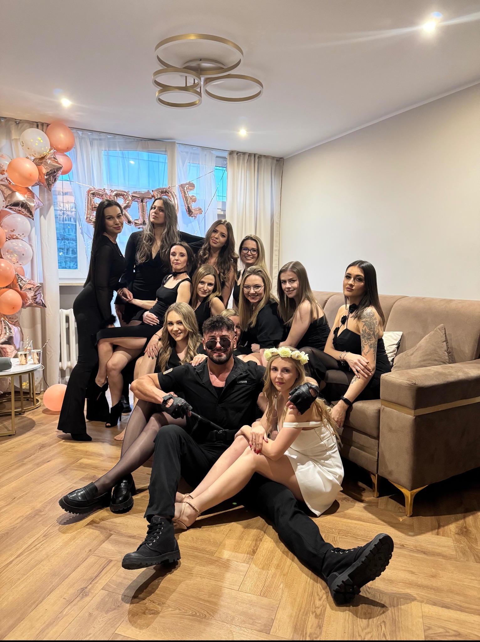 Wieczór panieński w mieszkaniu: grupa kobiet w czerni i mężczyzna w przebraniu siedzą na podłodze i sofie, dekoracje z balonów, napis 'Bride'.