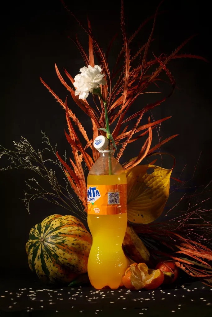 Martwa natura: butelka Fanta z kwiatem, dynia, mandarynki i suche gałęzie na ciemnym tle. Nietypowe połączenie elementów, miękkie światło.