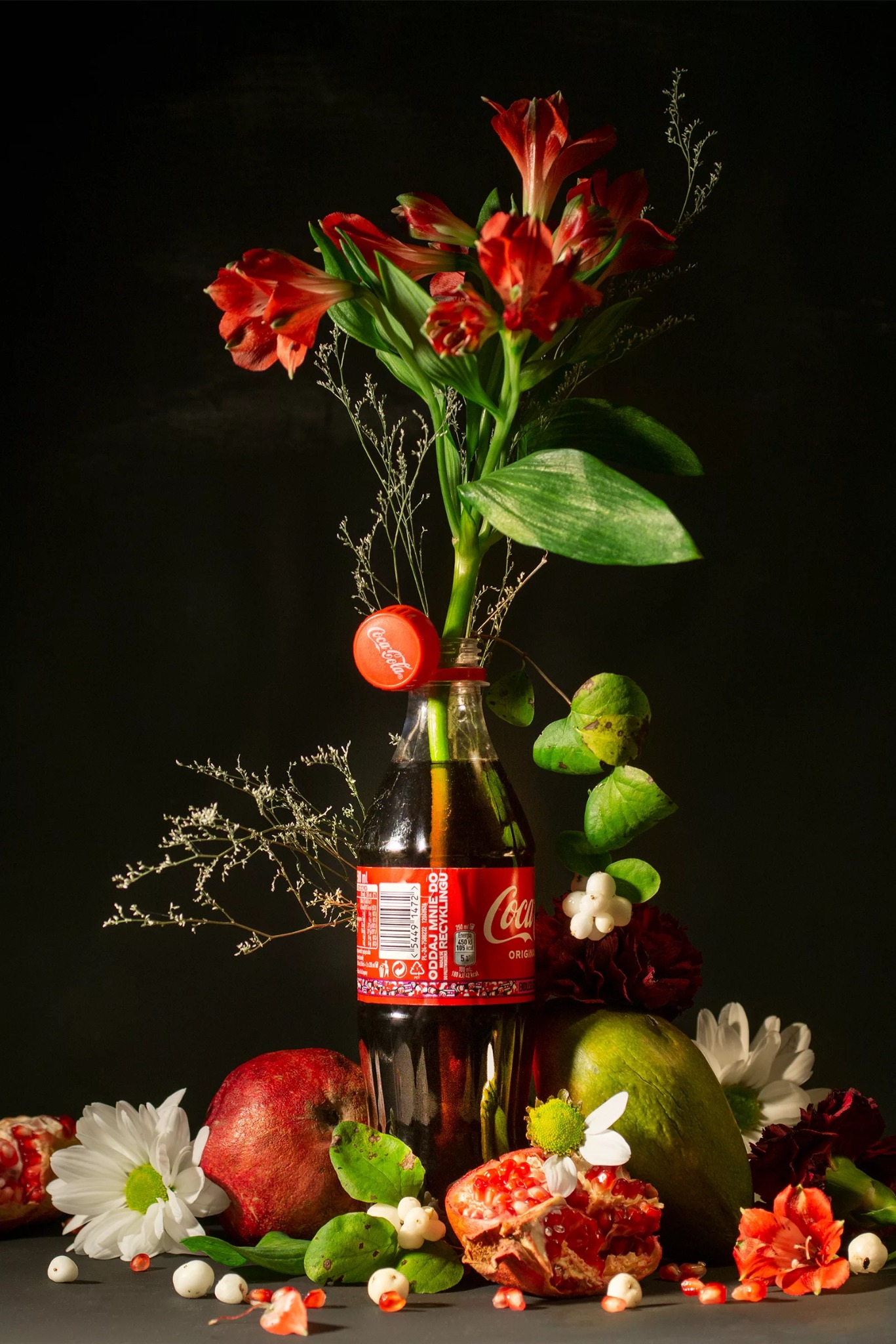 Martwa natura: Coca-Cola w butelce jako wazon z czerwonymi kwiatami i owocami granatu na ciemnym tle. Kompozycja z elementami florystycznymi i owocowymi.