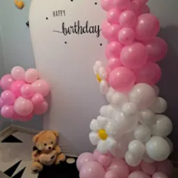 Dekoracja urodzinowa: białe tło z napisem 'Happy Birthday', girlanda z różowych i białych balonów w kształcie kwiatów, pluszowy miś na okrągłym dywanie.