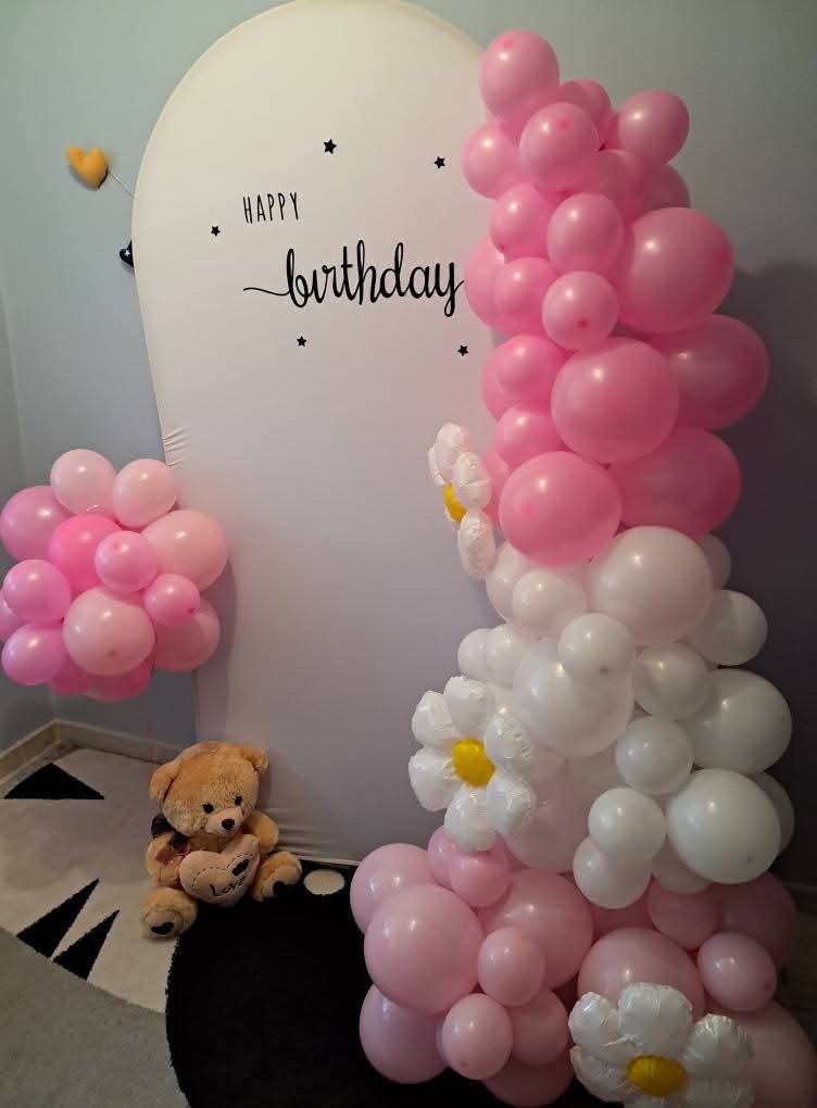 Dekoracja urodzinowa: białe tło z napisem 'Happy Birthday', girlanda z różowych i białych balonów w kształcie kwiatów, pluszowy miś na okrągłym dywanie.