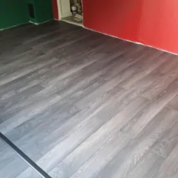 Ułożona podłoga z paneli w odcieniach szarości, ściany pomalowane na czerwono i zielono, listwy przypodłogowe w trakcie montażu. Widok z góry, ukośnie.