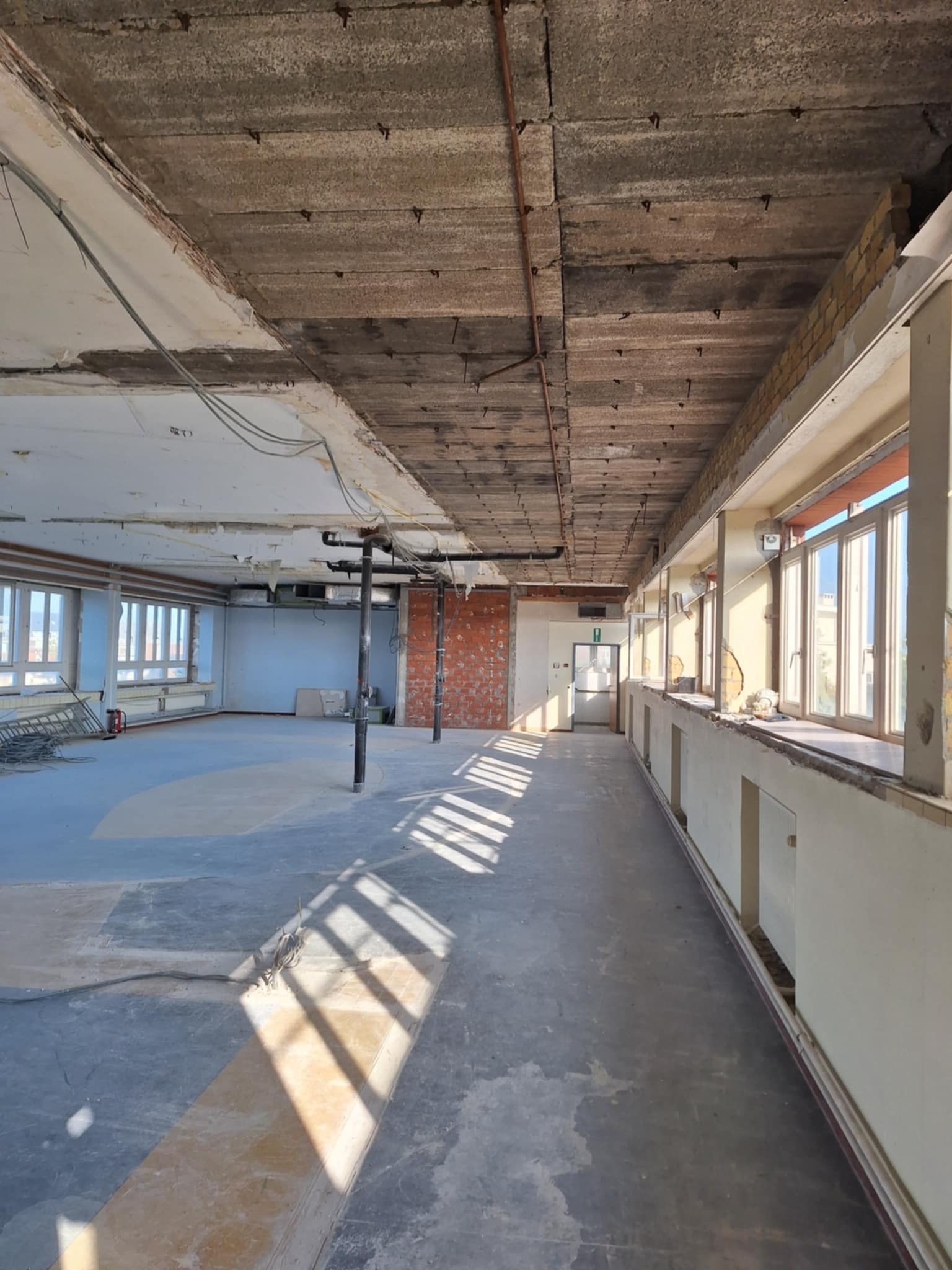 Wnętrze w trakcie rozbiórki: odsłonięty sufit z betonowych płyt, fragmenty ścian z cegły, okna z widokiem na miasto, betonowa podłoga z resztkami wykładziny.
