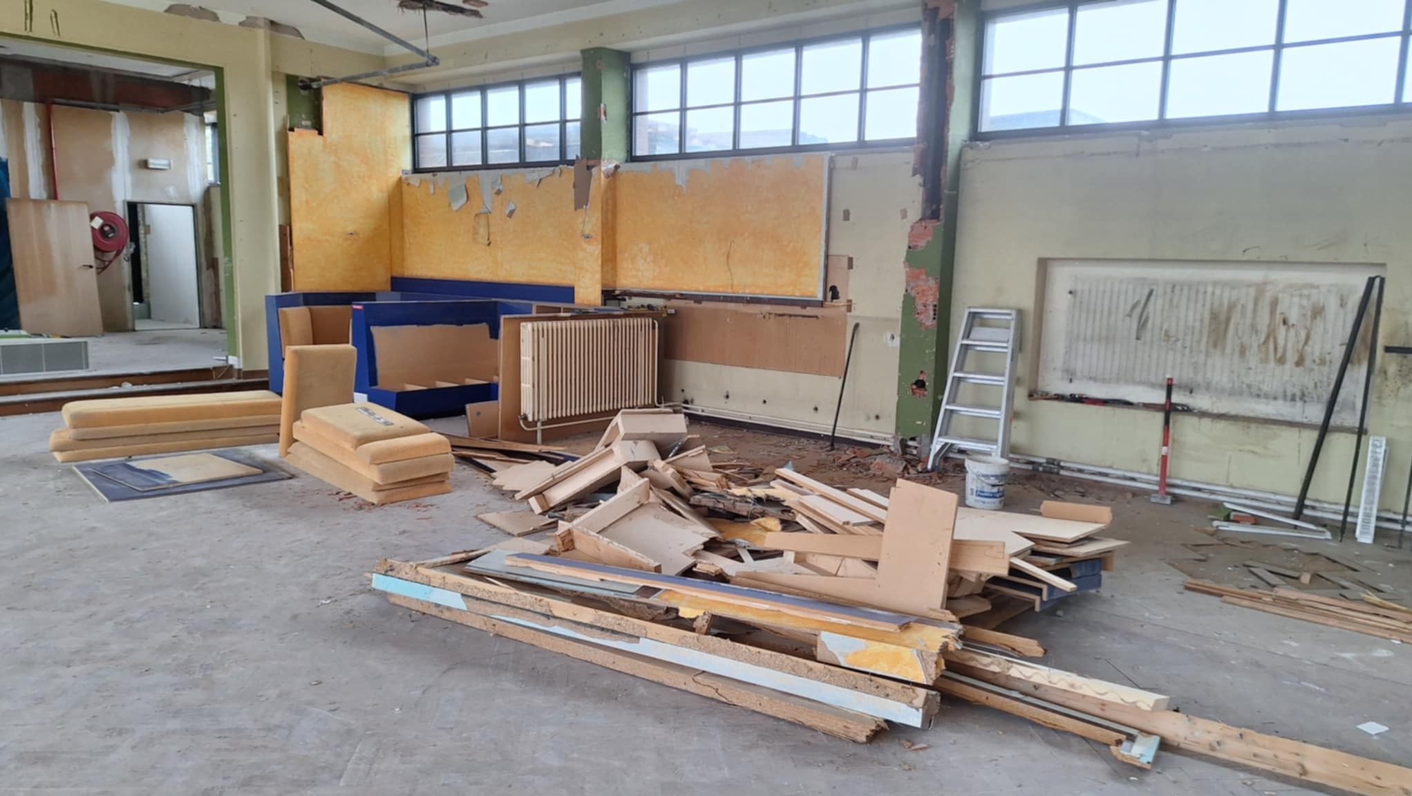 Wnętrze po rozbiórce: gruz, resztki mebli i ściany z odsłoniętą cegłą. Widoczne schody i drabina. Duże okna w tle. Pomieszczenie w trakcie prac rozbiórkowych.
