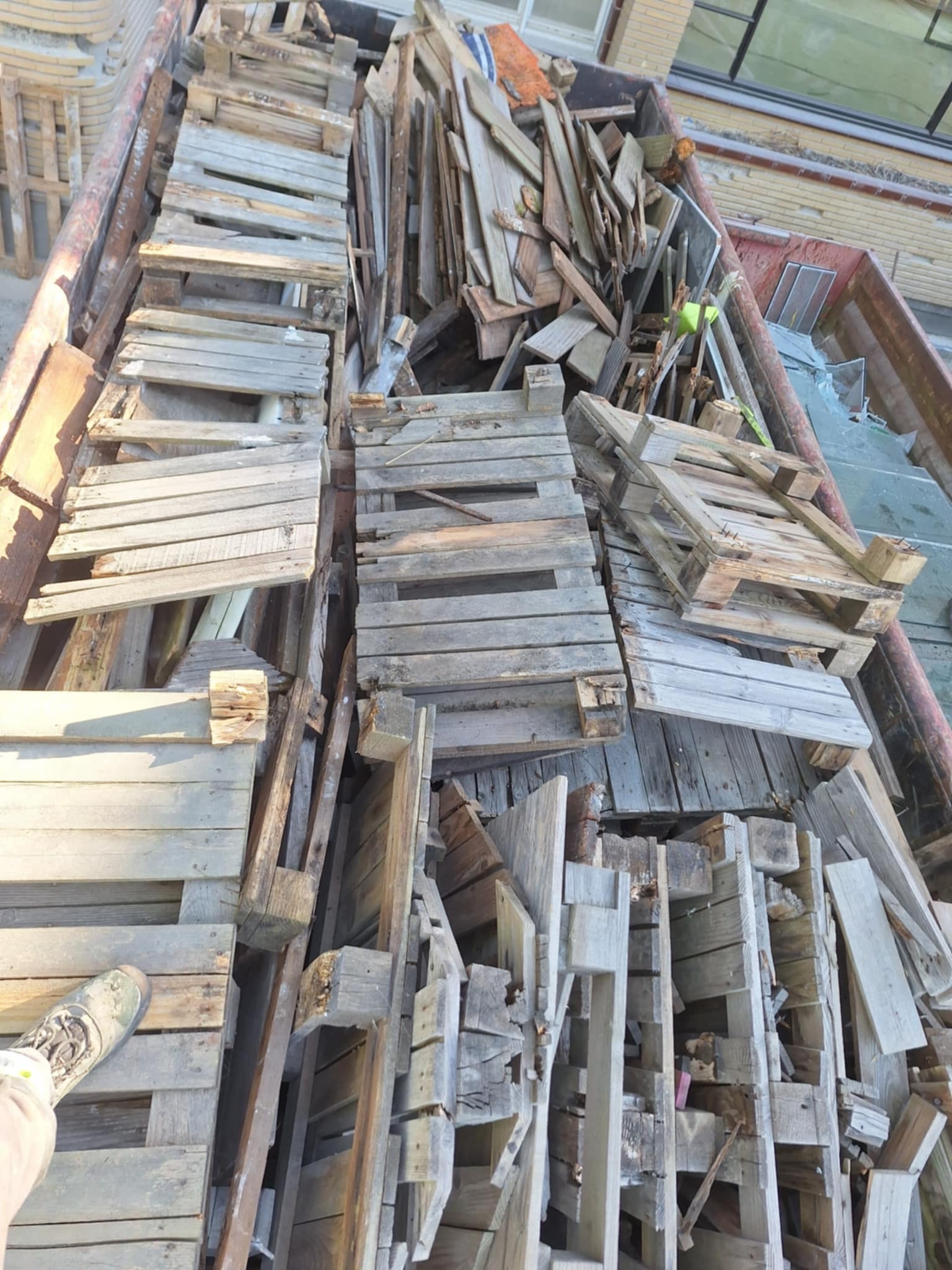 Kontener wypełniony starymi, drewnianymi paletami i fragmentami desek po rozbiórce, z widocznym fragmentem buta osoby stojącej obok.