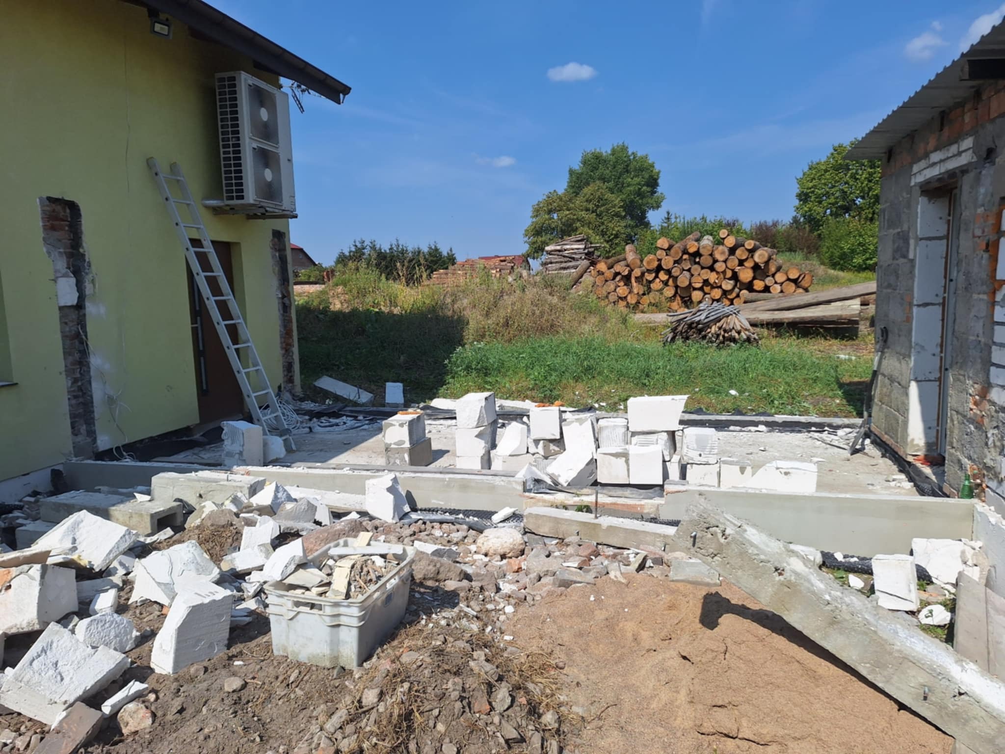 Po rozbiórce: fundament domu z pustaków, gruz, piasek. W tle sterta drewna i budynek w trakcie demontażu. Drabina oparta o ścianę budynku.