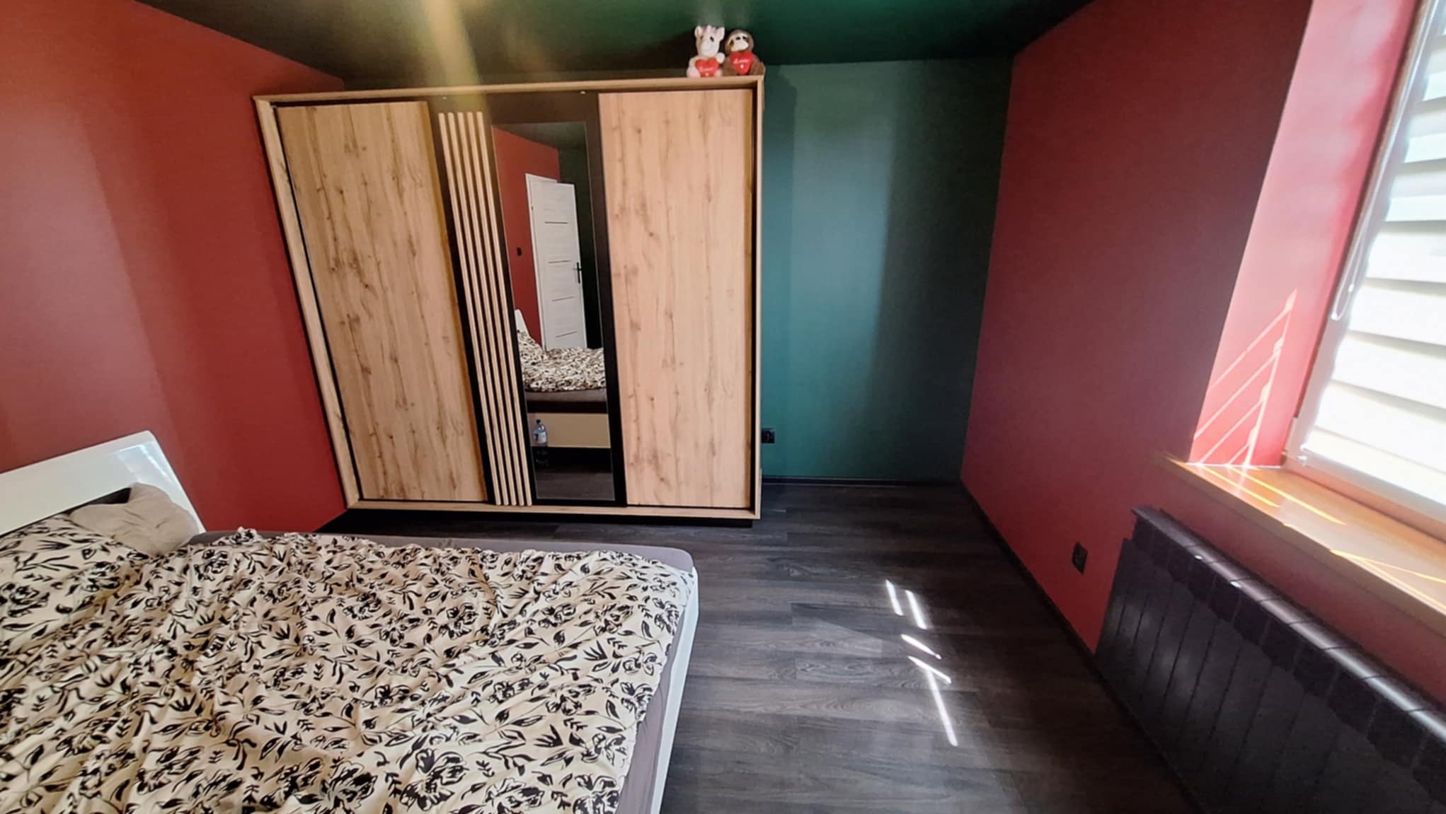 Sypialnia po remoncie: łóżko z wzorzystą pościelą, drewniana szafa z lustrem, ściany w odcieniach czerwieni i zieleni, ciemna podłoga. Pluszaki na szafie.
