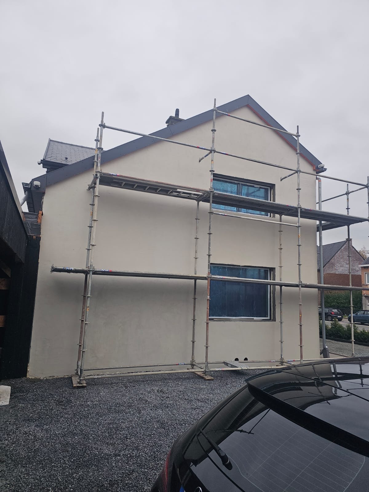 Otynkowany dom jednorodzinny w trakcie prac elewacyjnych, z widocznymi rusztowaniami i zabezpieczonymi oknami. Szara dachówka i elewacja w odcieniach beżu.