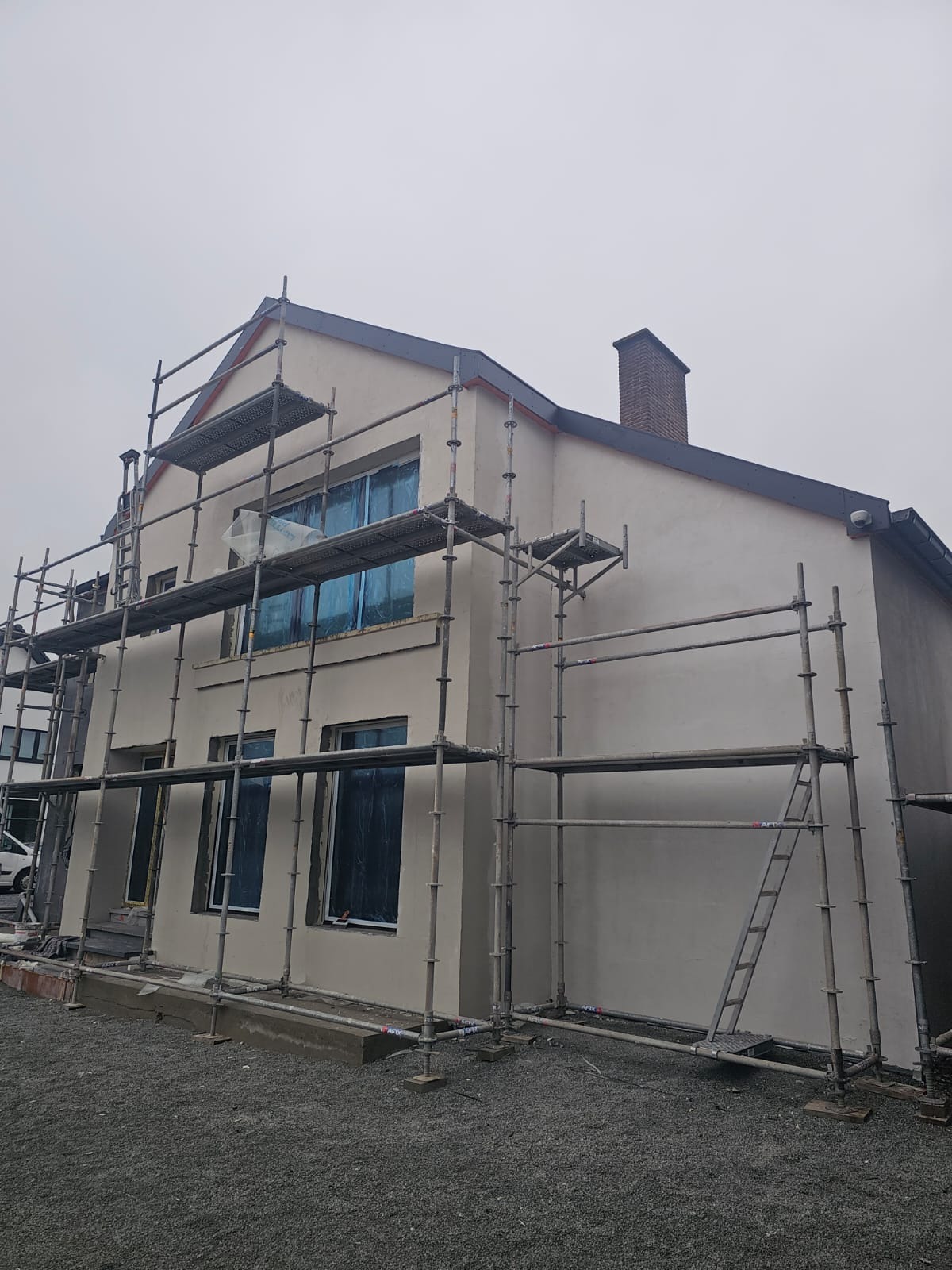 Dom w trakcie ocieplania, osłonięte okna folią, widoczne rusztowania. Świeżo nałożony tynk w kolorze beżowym. Kompozycja z dołu, pochmurne niebo.