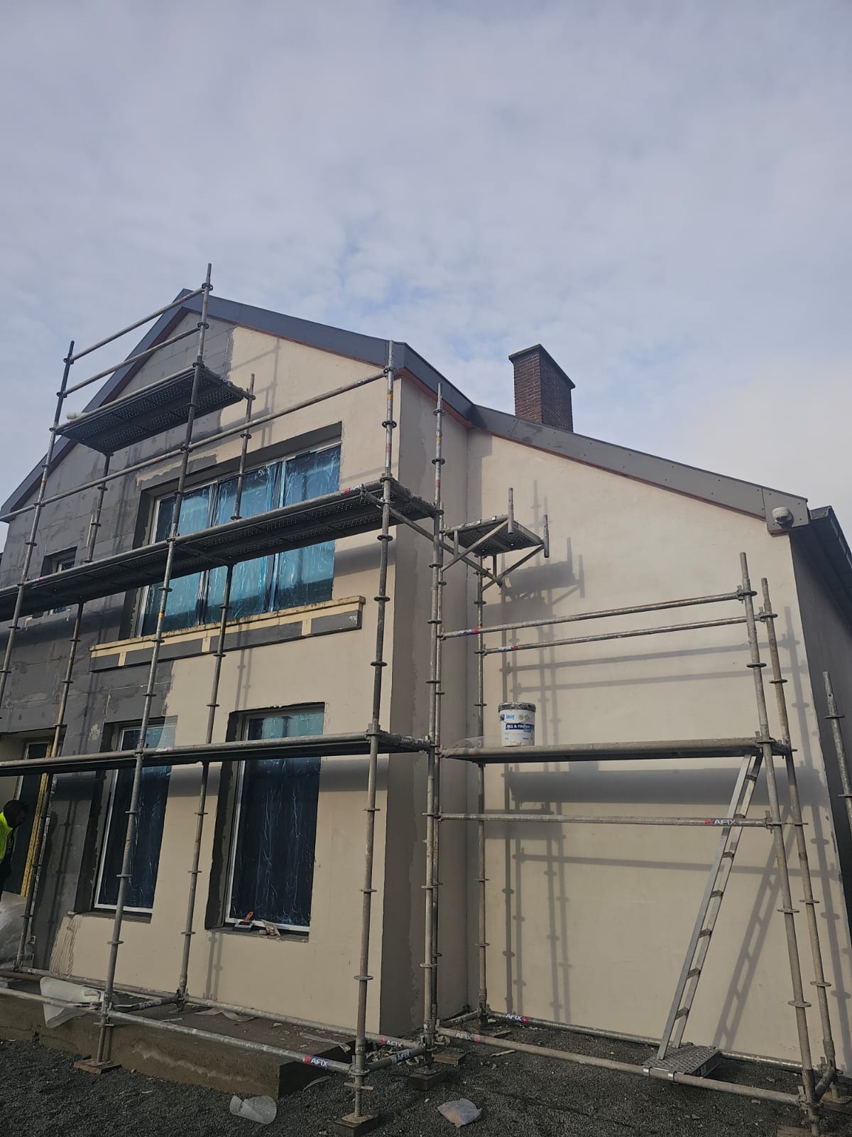 Ocieplanie elewacji domu jednorodzinnego w trakcie realizacji. Widoczne rusztowania, zabezpieczone okna folią i fragment ocieplonej ściany w kolorze szarym.