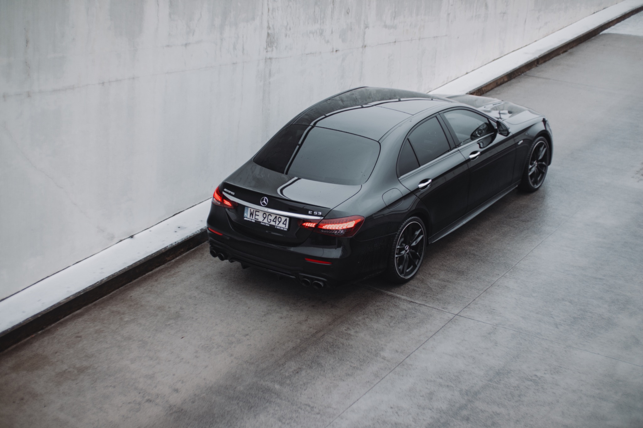 Czarny Mercedes-AMG E 53, widok z góry na tle betonowej ściany. Widoczne detale: światła, felgi, tablica rejestracyjna. Minimalistyczna kompozycja, stonowana kolorystyka.