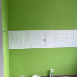 Michał Wilk - Ściana pomalowana na zielono z pasem białych płytek i gniazdkiem. Na podłodze narzędzia i materiały malarskie: pędzle, wiadro z farbą, butelka z wodą i pistolet do silikonu.