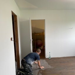 Michał Wilk - Mężczyzna na kolanach czyści drewnianą podłogę gąbką w remontowanym pokoju z białymi ścianami. Widoczne otwarte drzwi i instalacja elektryczna.