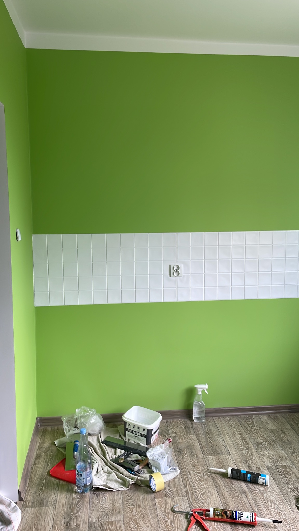 Ściana pomalowana na zielono z pasem białych płytek i gniazdkiem. Na podłodze narzędzia i materiały malarskie: pędzle, wiadro z farbą, butelka z wodą i pistolet do silikonu.