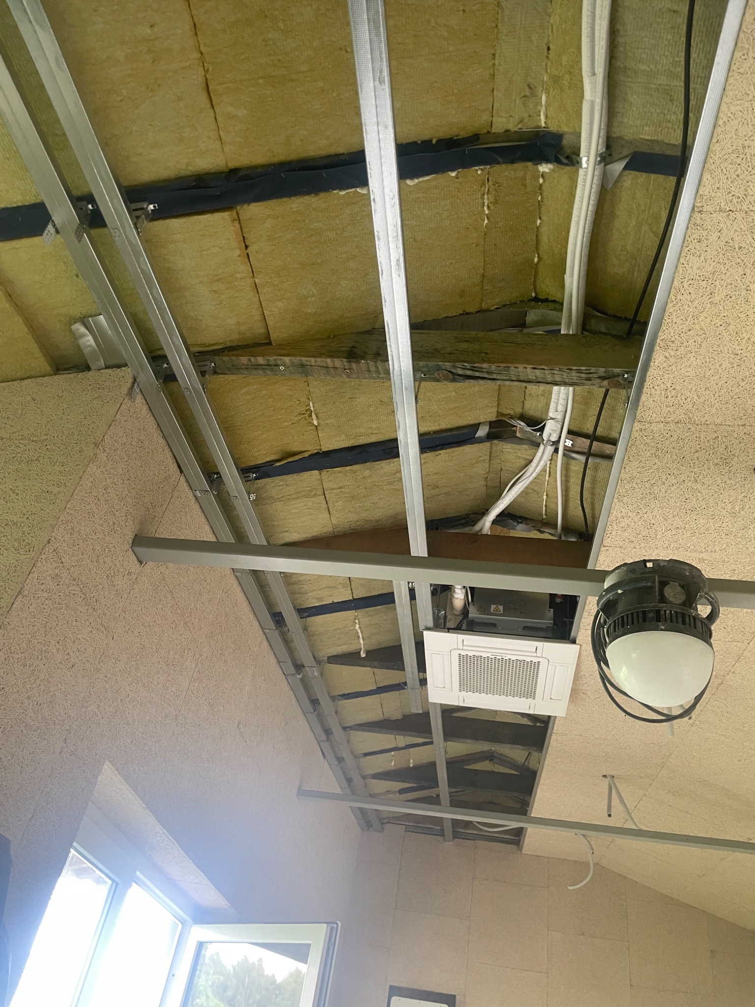 Skośny sufit z izolacją z wełny mineralnej, metalowym stelażem i płytami OSB. Widoczna klimatyzacja i oświetlenie. Ujęcie z dołu, okno w tle.
