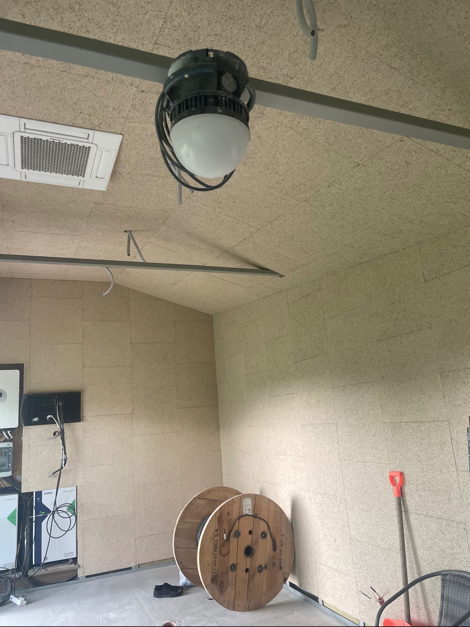 Wnętrze pomieszczenia z płytami OSB na ścianach i suficie, widoczna lampa przemysłowa, klimatyzacja, przewody elektryczne i bębny z kablem, sugerujące etap prac wykończeniowych.