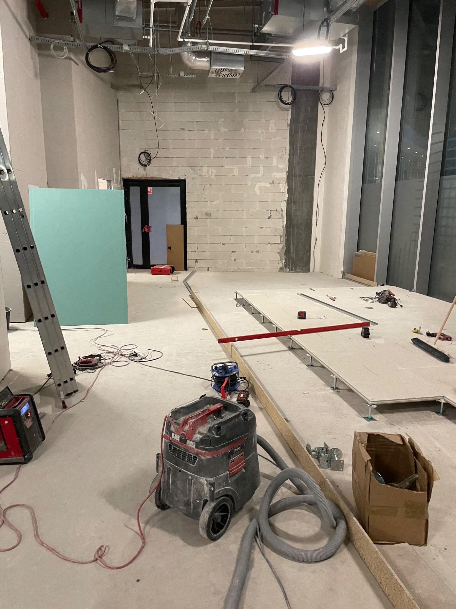 Wnętrze w trakcie prac wykończeniowych: surowe ściany, podłoga z płyt OSB na regulowanych wspornikach, drabina, narzędzia i odkurzacz przemysłowy. Widoczne instalacje pod sufitem.