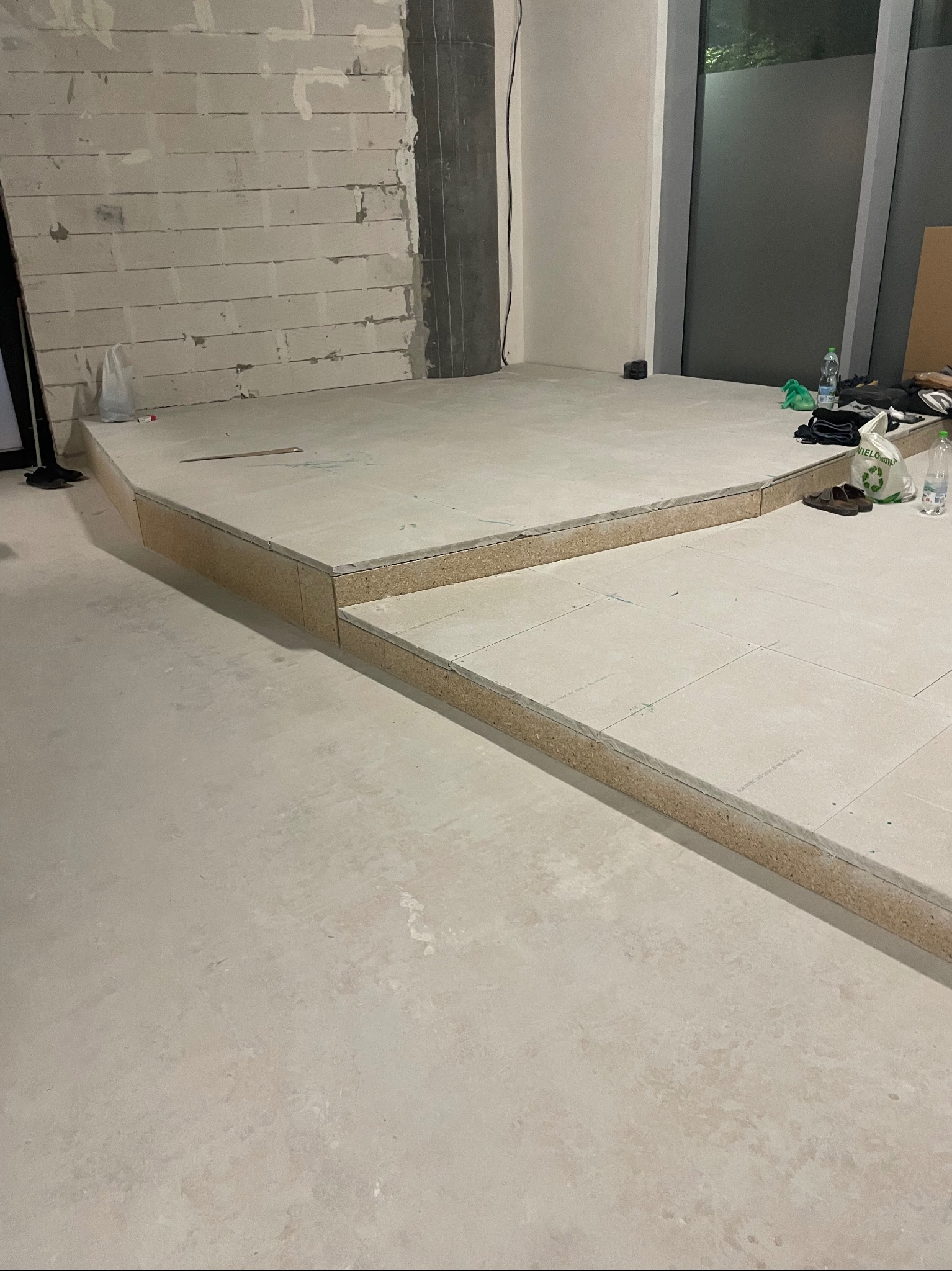 Wnętrze w trakcie prac wykończeniowych: podwyższona podłoga z płyt, surowa ściana z widocznymi spoinami, duże okno. Widoczne materiały budowlane i akcesoria.