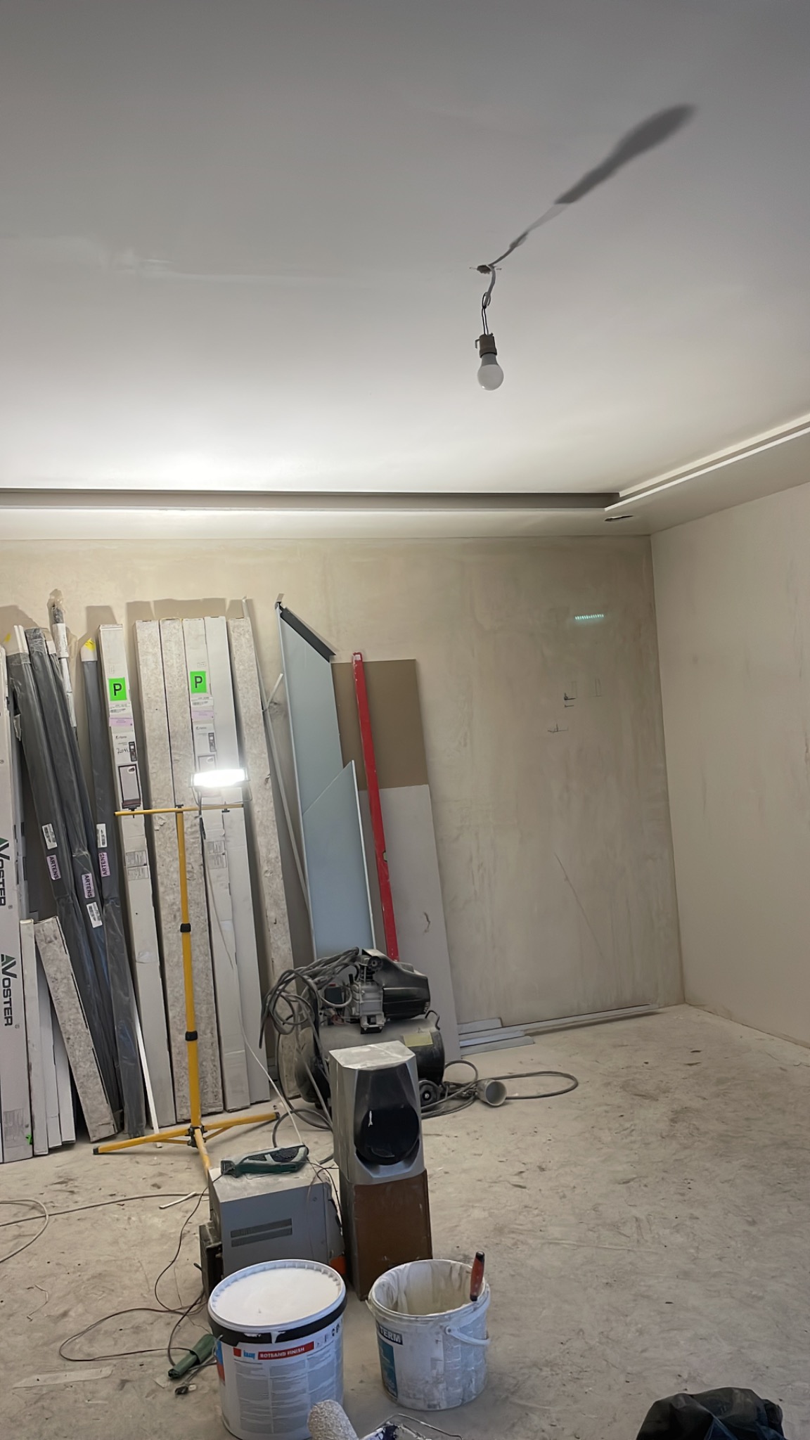 Przygotowania do malowania ścian w pustym pomieszczeniu, widoczne narzędzia, pędzle i farby, sufit z żarówką na kablu, płyty karton-gips oparte o ścianę.