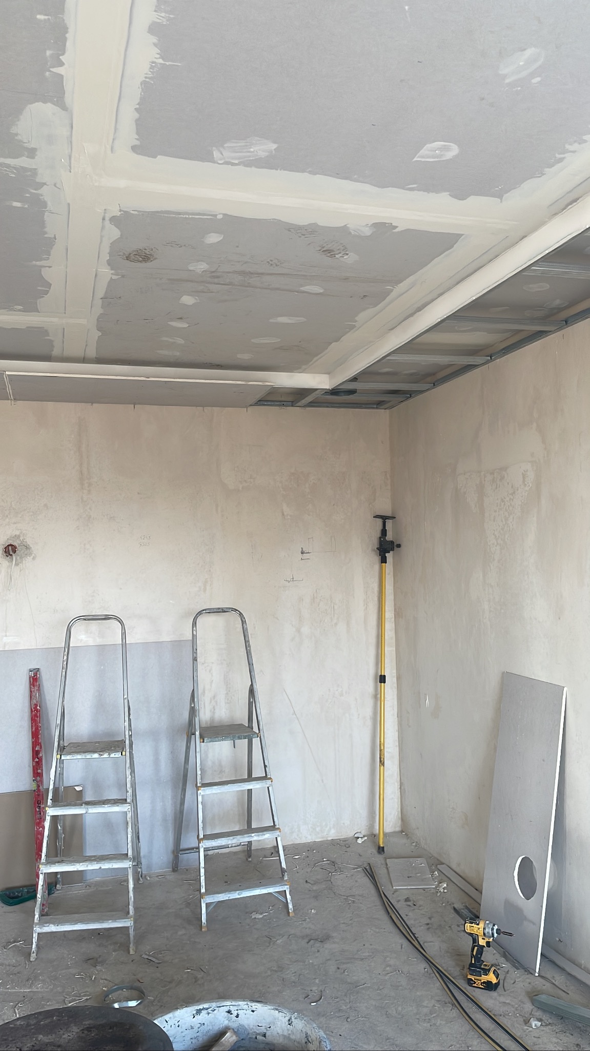Sufit z płyt gipsowo-kartonowych w trakcie szpachlowania, widoczne metalowe profile, drabiny i narzędzia w surowym pomieszczeniu. Etap prac wykończeniowych.