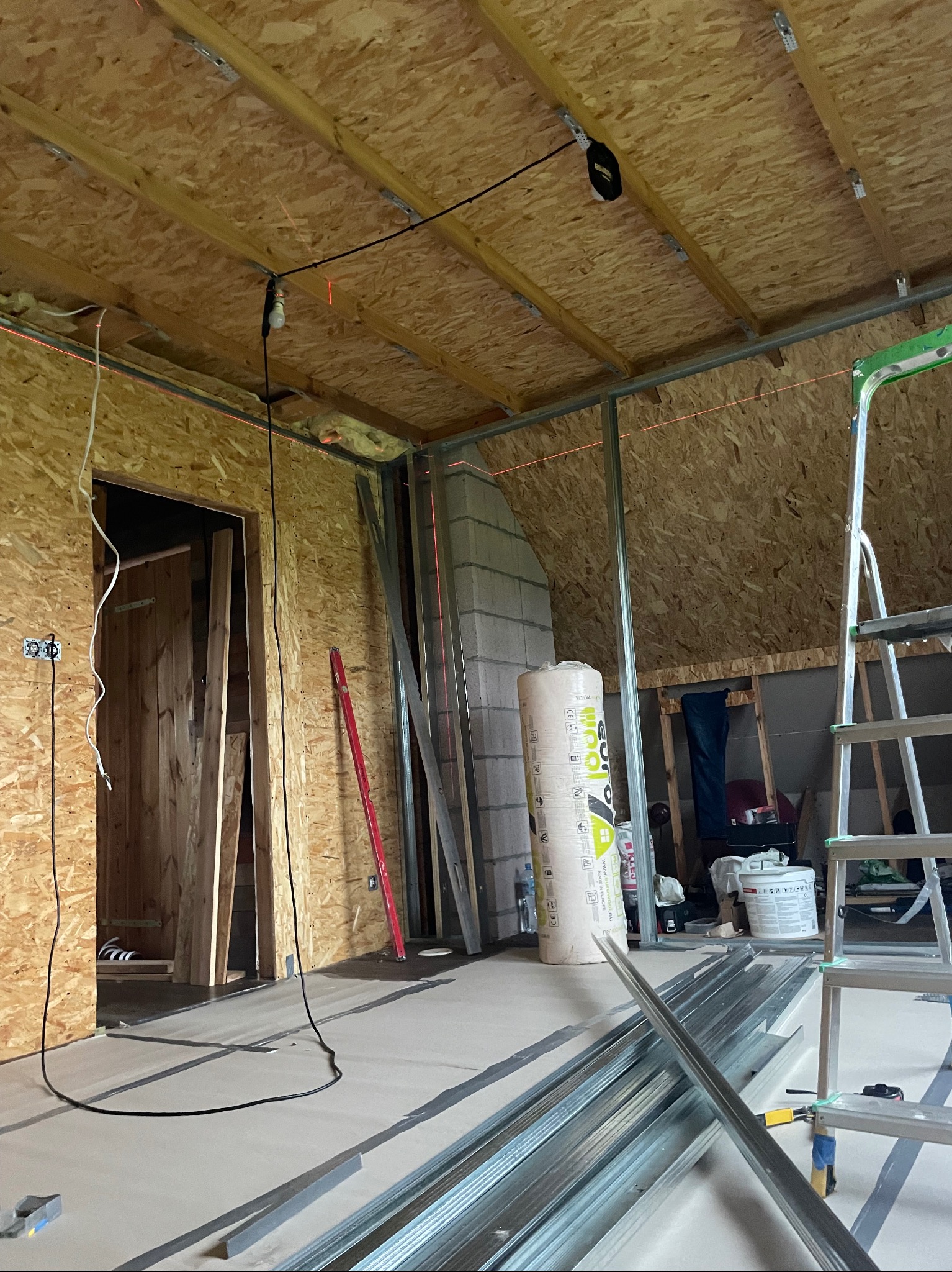 Poddasze w trakcie adaptacji: ściany obite płytą OSB, metalowe profile, izolacja, poziomica laserowa. Widoczna drabina i narzędzia. Prace wykończeniowe.
