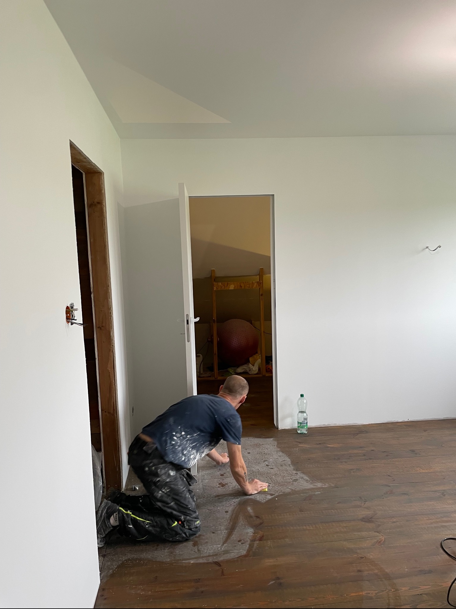 Mężczyzna na kolanach czyści drewnianą podłogę gąbką w remontowanym pokoju z białymi ścianami. Widoczne otwarte drzwi i instalacja elektryczna.
