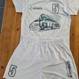 Biały komplet sportowy (koszulka i spodenki) z nadrukiem 'VOLLEYBALL', numerem '5' i logo 'KOMBUD', leżący na drewnianym stole. Widoczne detale nadruku.