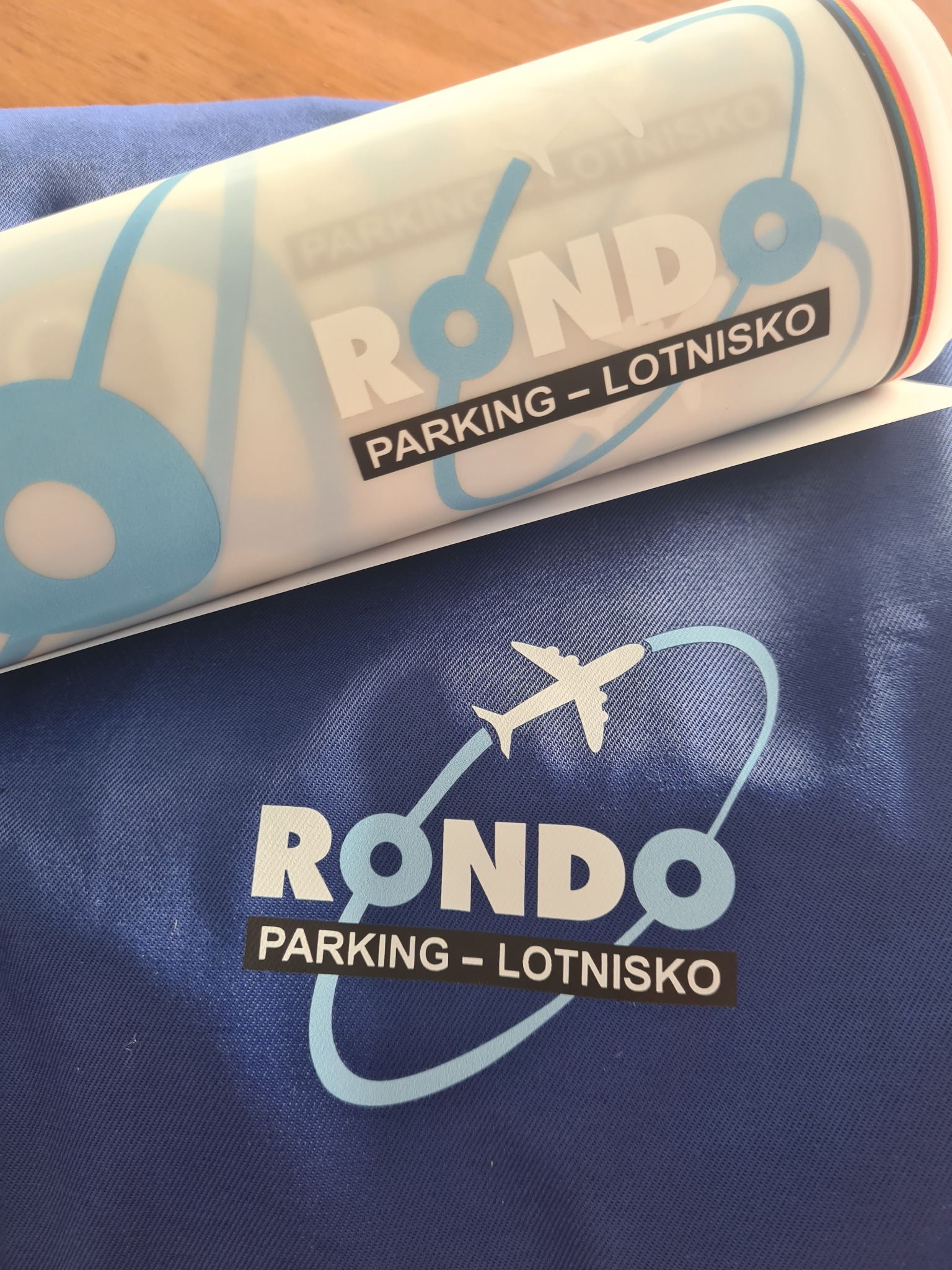Niebieska torba z nadrukiem logo 'RONDO PARKING - LOTNISKO' i szablon do nadruku z tym samym wzorem, leżące na drewnianym stole.