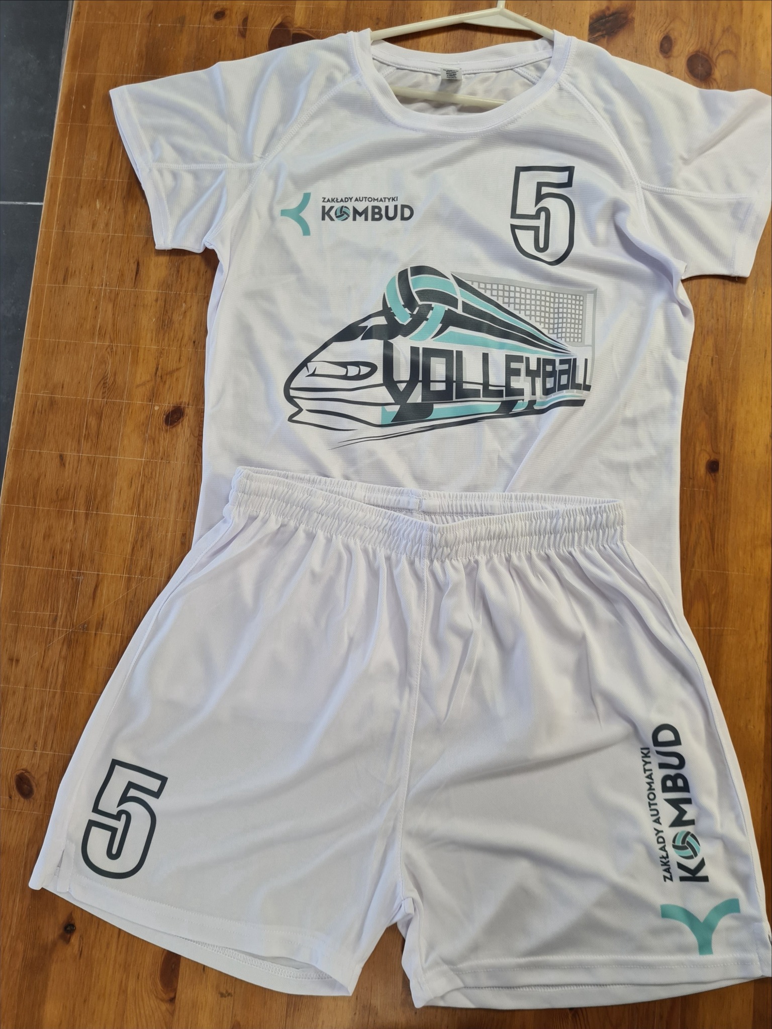 Biały komplet sportowy (koszulka i spodenki) z nadrukiem 'VOLLEYBALL', numerem '5' i logo 'KOMBUD', leżący na drewnianym stole. Widoczne detale nadruku.