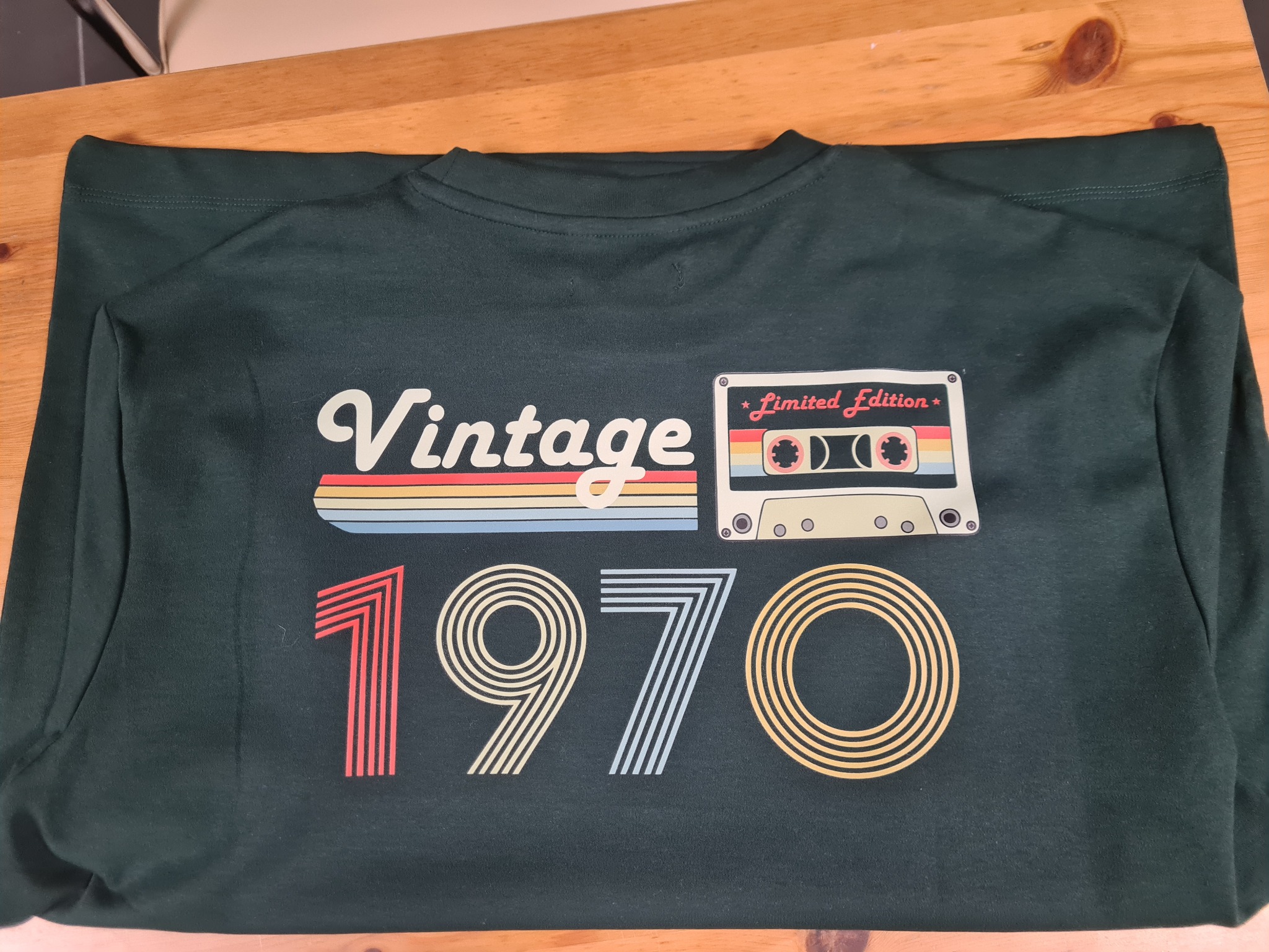 Ciemnozielona koszulka z nadrukiem 'Vintage 1970' i motywem kasety magnetofonowej leży na drewnianym stole. Grafika w stylu retro.