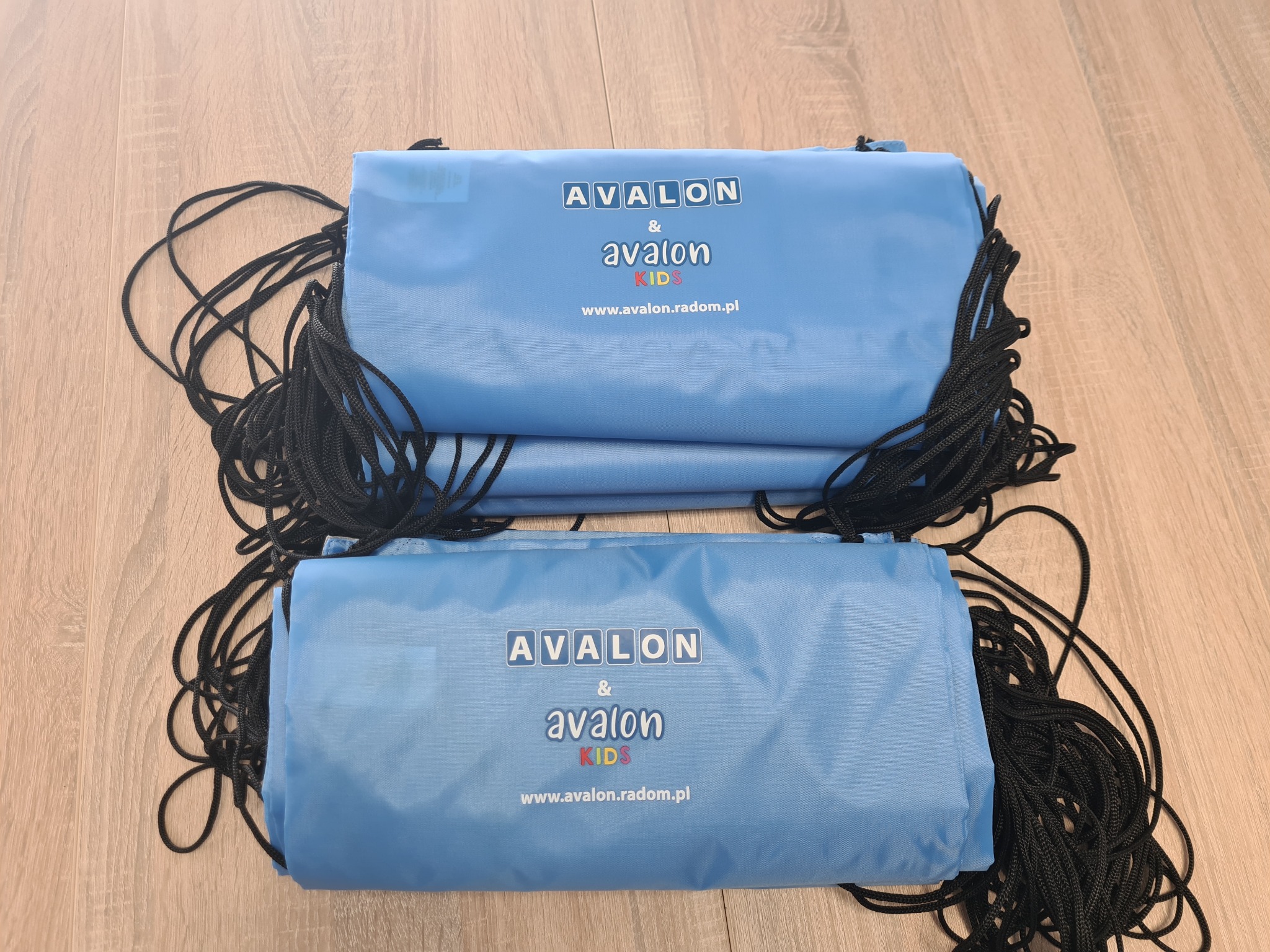 Dwa niebieskie worki z nadrukiem 'AVALON & avalon KIDS' i czarnymi sznurkami, leżące na drewnianej podłodze. Widoczne logo firmy i adres strony internetowej.