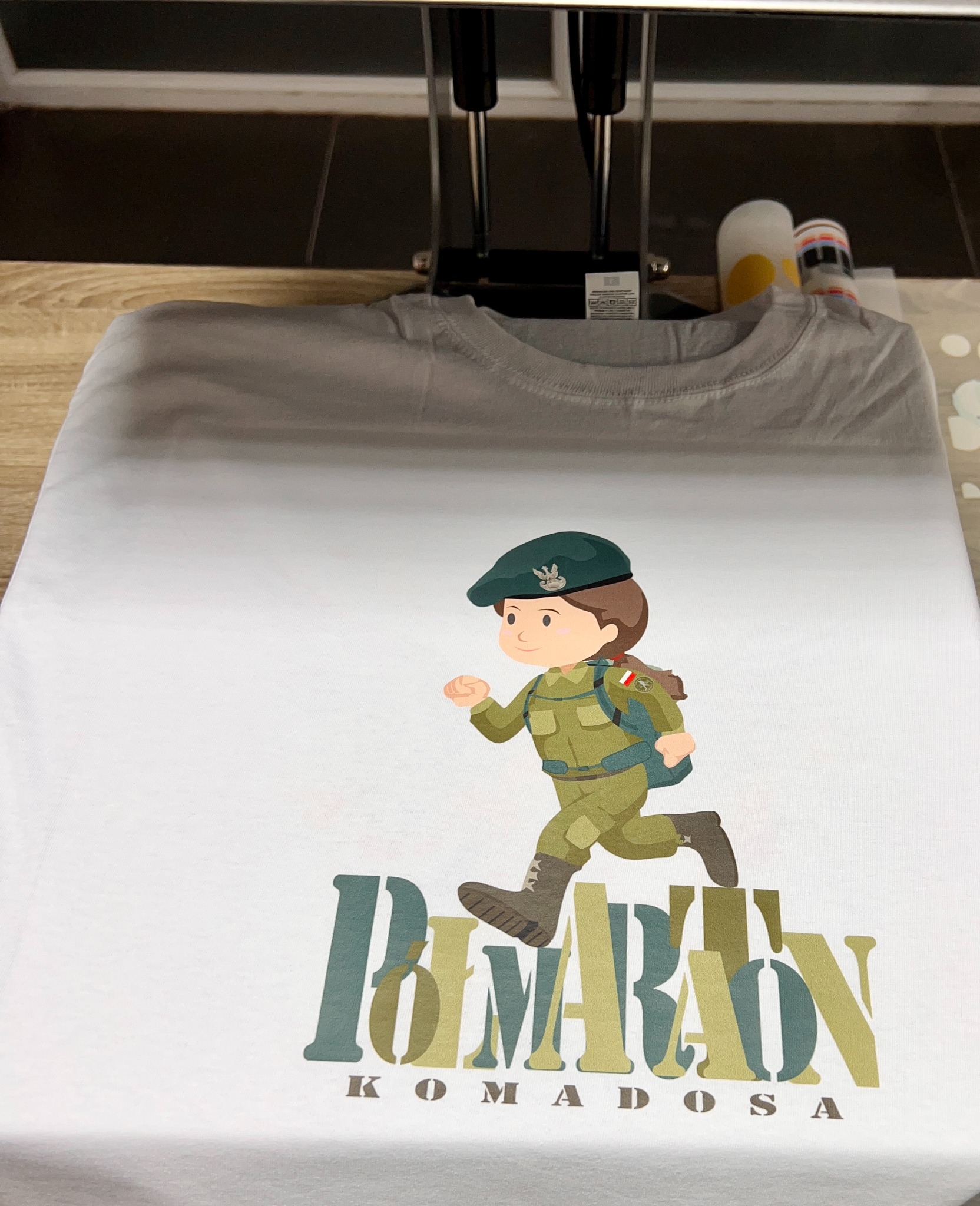 Biała koszulka z nadrukiem: postać w mundurze i napis 'BIMARDON KOMADOSA'. Widoczna prasa termotransferowa. Gotowy produkt po wykonaniu usługi.