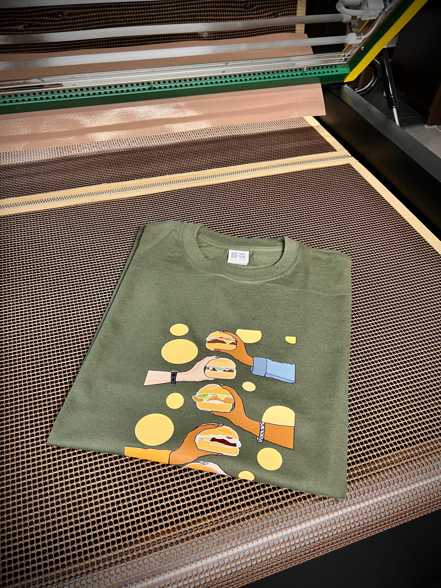 Oliwkowa koszulka z nadrukiem rąk trzymających burgery, leżąca na perforowanym stole w warsztacie.