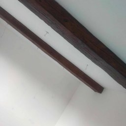 JARBUD FIRMA OGÓLNOBUDOWLANA JAROSŁAW JANUSZKIEWICZ - Białe wnętrze z dwiema ciemnymi, drewnianymi belkami stropowymi, zawieszonymi na metalowych haczykach, widocznymi pod kątem od dołu.