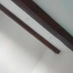 Białe wnętrze z dwiema ciemnymi, drewnianymi belkami stropowymi, zawieszonymi na metalowych haczykach, widocznymi pod kątem od dołu.