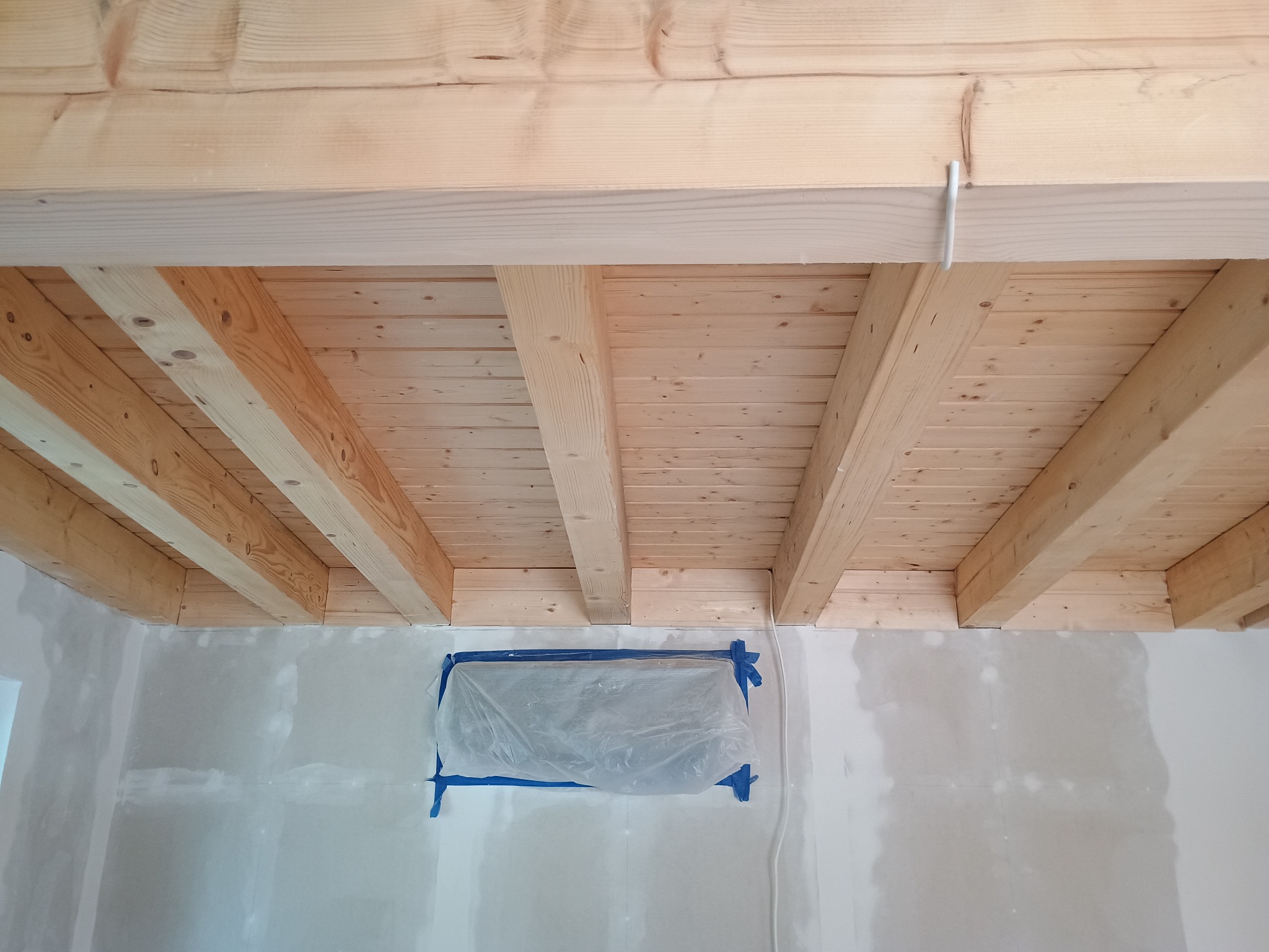 Sufit z drewnianych belek i ściana po szpachlowaniu, okno zabezpieczone folią. Widoczne ślady szpachli na ścianie wskazują na etap prac wykończeniowych.