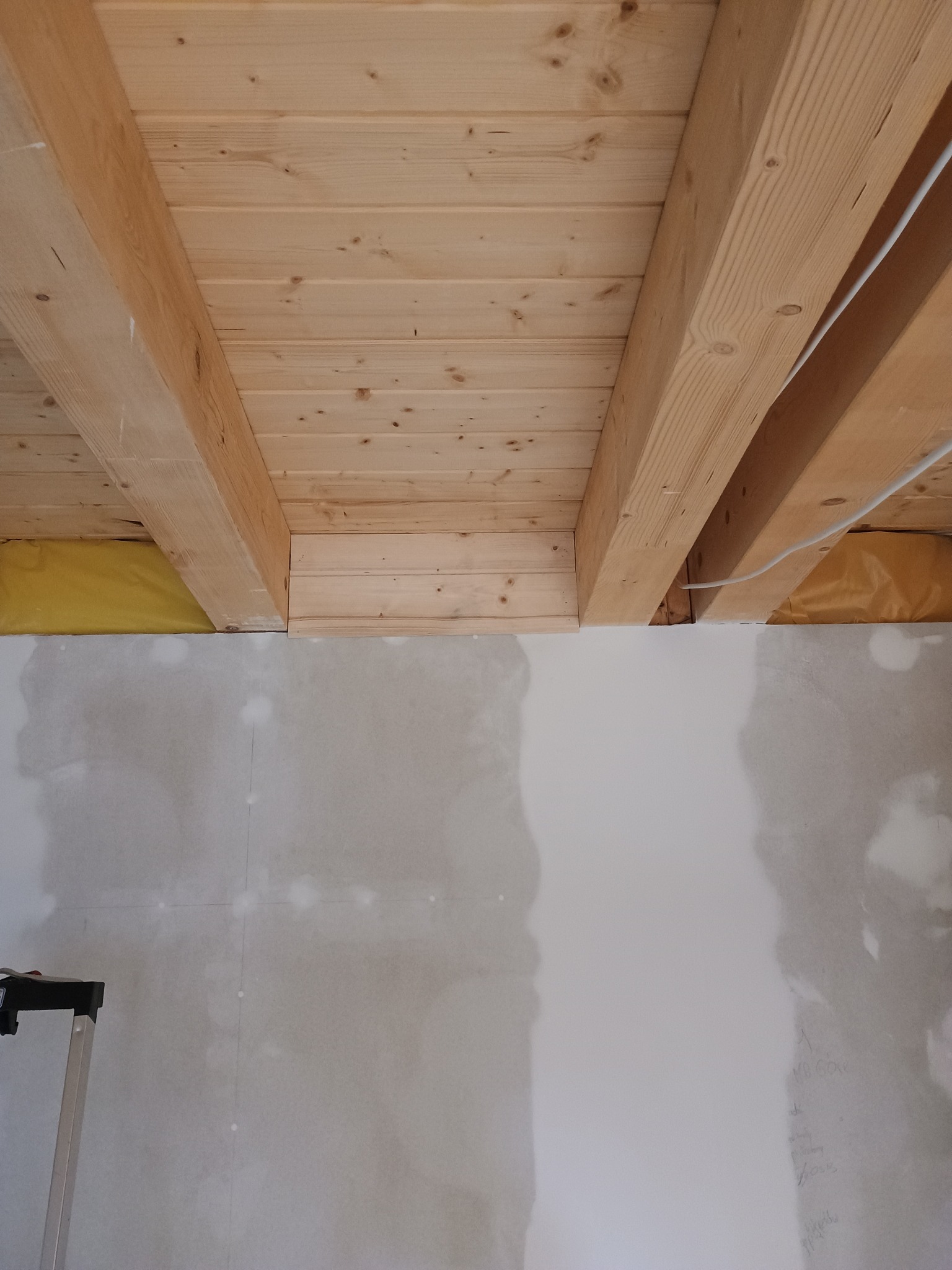 Sufit z belek drewnianych i fragment ściany po szpachlowaniu, widoczne żółte ocieplenie. Ujęcie od dołu, widać niedoskonałości powierzchni ściany.