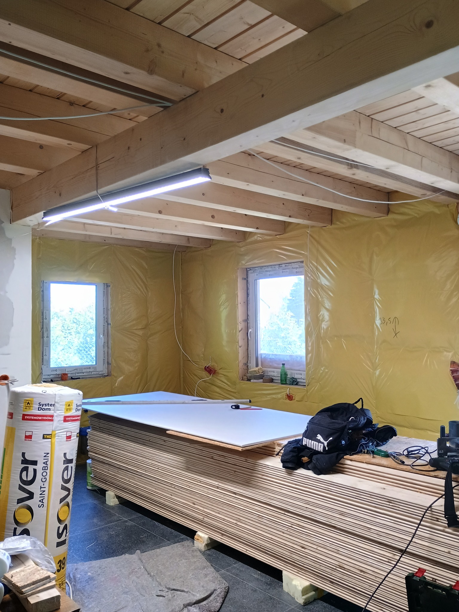 Wnętrze w trakcie prac wykończeniowych: ocieplone ściany folią, drewniany sufit z oświetleniem LED, stos płyt gipsowych i materiały izolacyjne Isover.