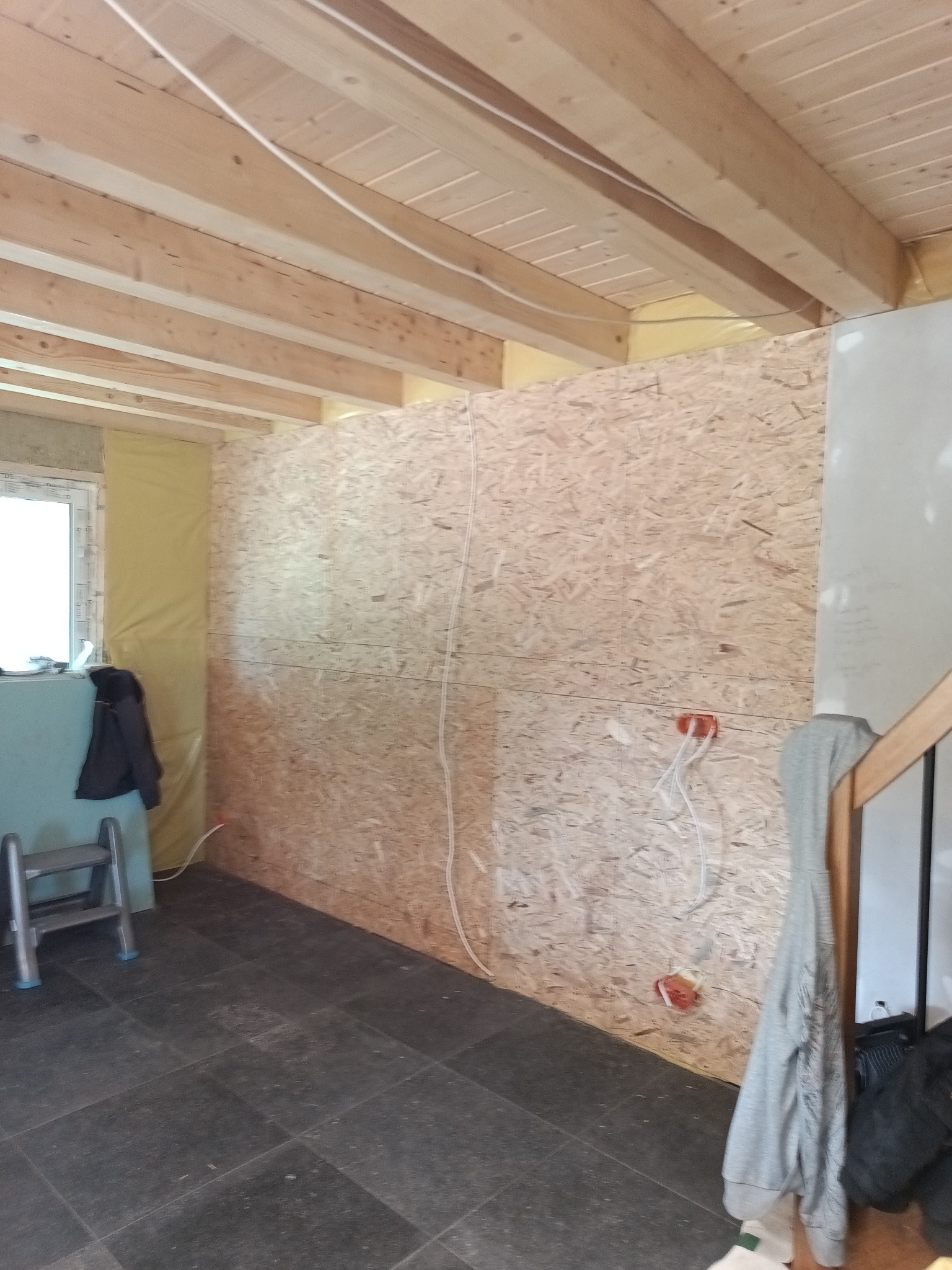 Wnętrze w trakcie prac wykończeniowych: ściany z płyt OSB z widoczną instalacją elektryczną, drewniany strop, fragment schodów i podłoga z ciemnych płytek.