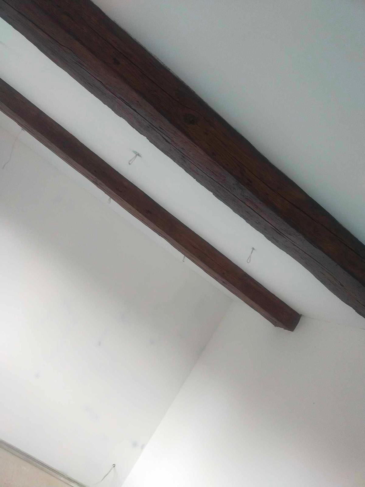 Białe wnętrze z dwiema ciemnymi, drewnianymi belkami stropowymi, zawieszonymi na metalowych haczykach, widocznymi pod kątem od dołu.