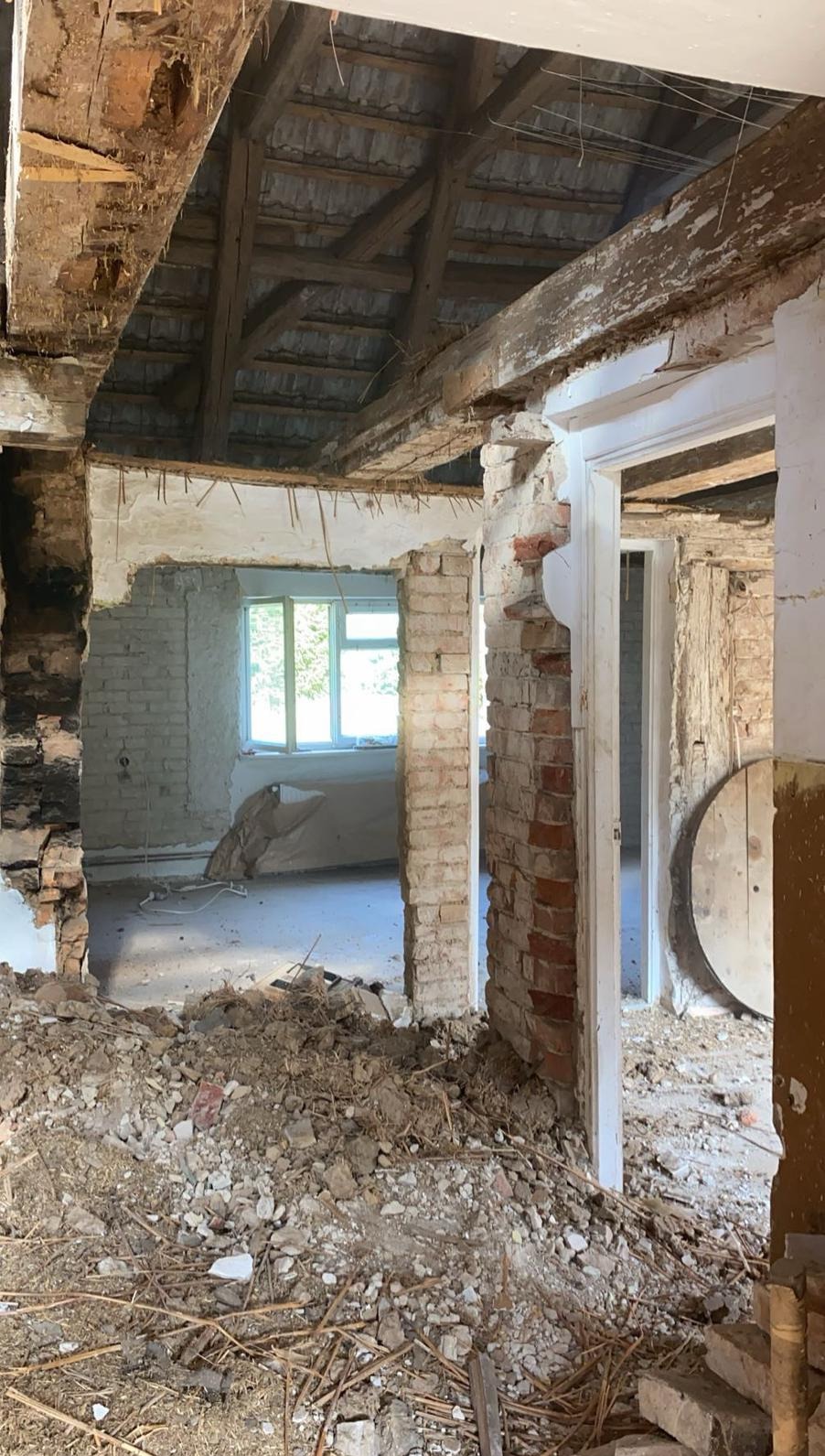 Demontaż ścian z cegły w trakcie remontu domu. Widoczne drewniane belki stropowe i gruz na podłodze. Okno z widokiem na zieleń w tle.