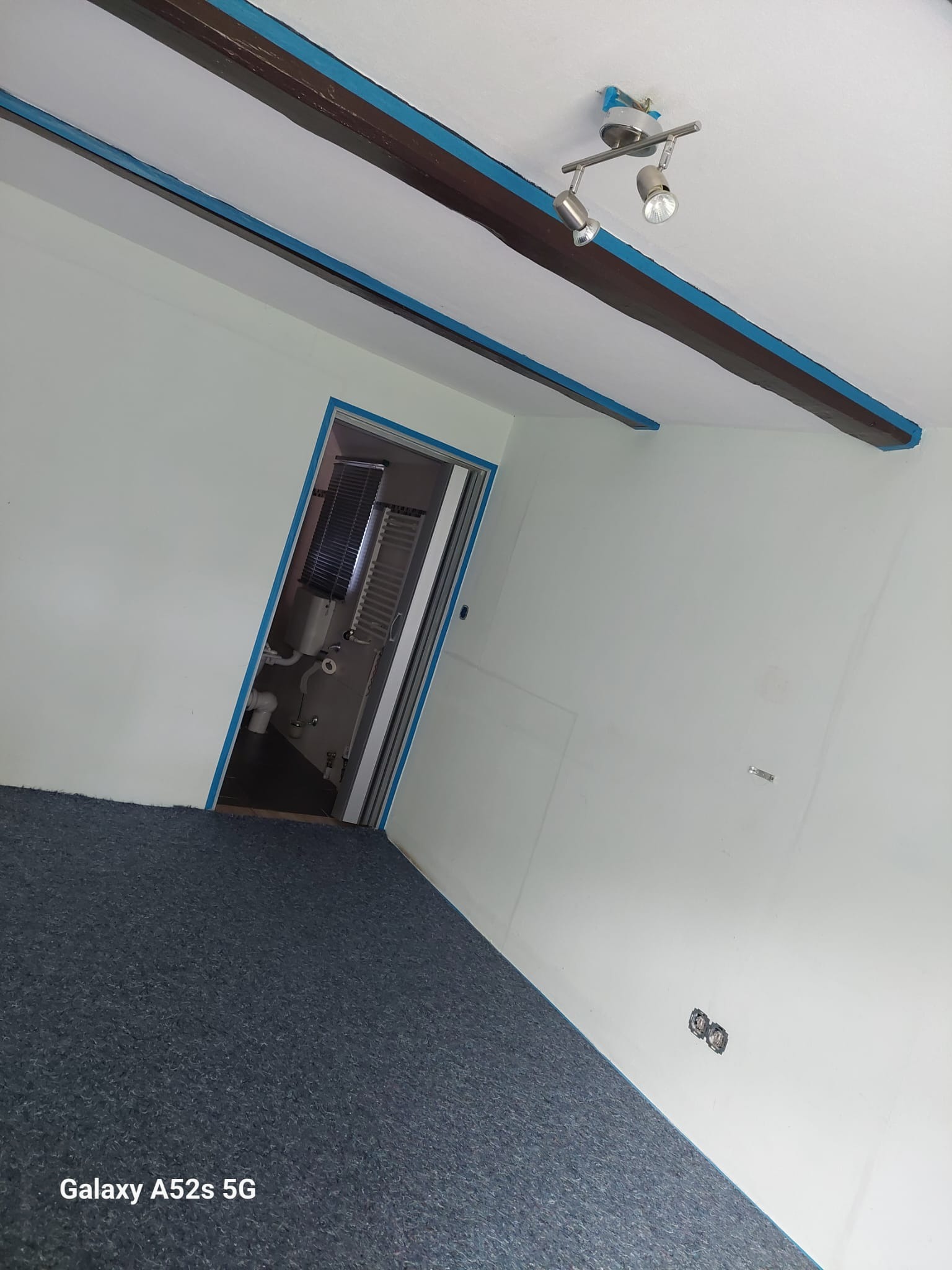 Białe ściany z drewnianymi belkami i niebieską taśmą malarską, widok na łazienkę, szara wykładzina. Przygotowanie do malowania.