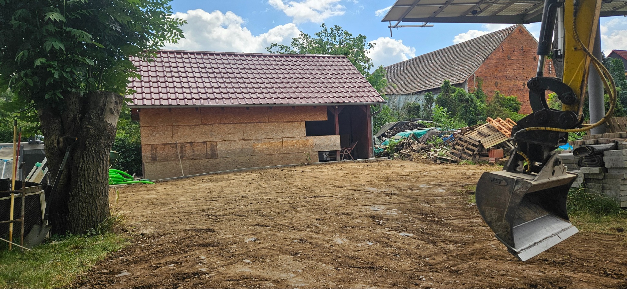 Przygotowany teren pod budowę z widoczną koparką i stertą materiałów budowlanych w tle. Widoczny budynek w trakcie budowy z płyt OSB.